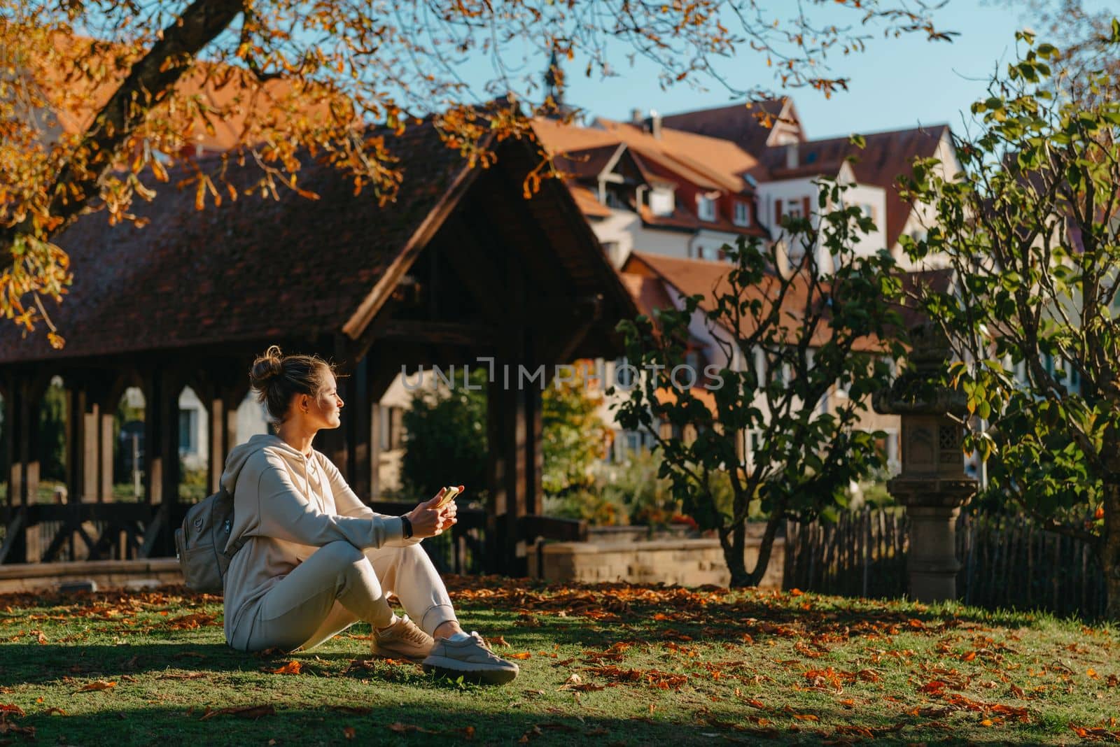 Young fashionable teenage girl with smartphone in park in autumn sitting at smiling. Trendy young woman in fall in park texting. Retouched, vibrant colors. Beautiful blonde teenage girl wearing casual modern autumn outfit sitting in park in autumn. Retouched, vibrant colors, brownish tones.