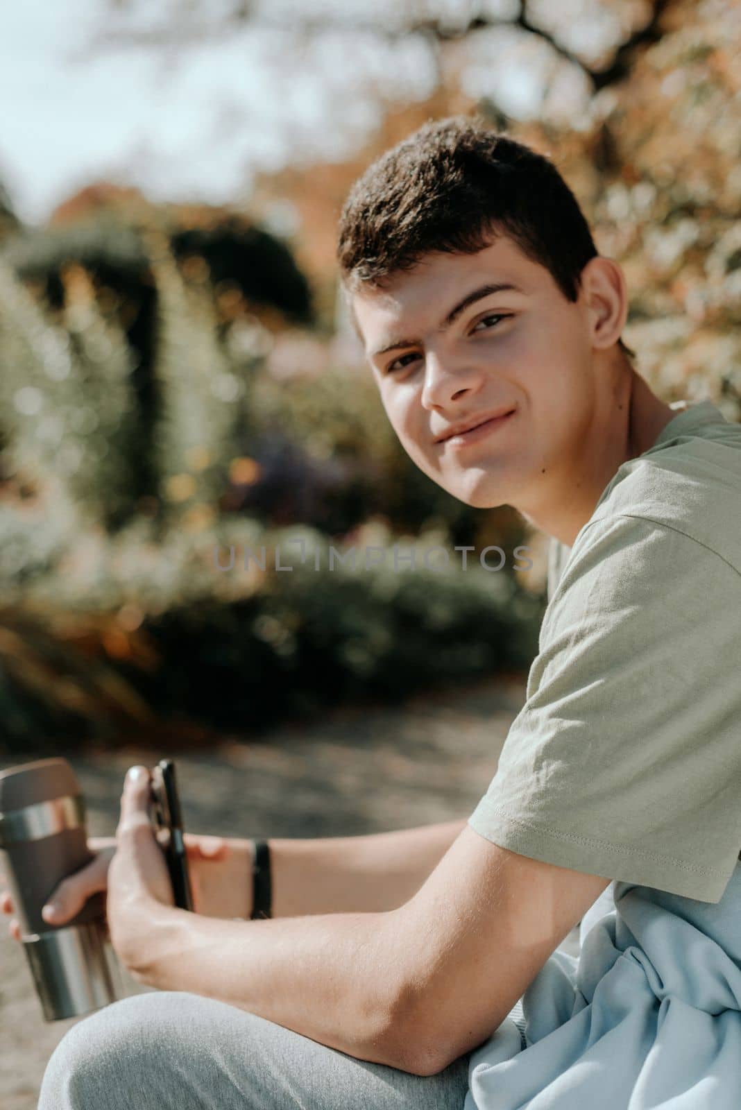 A Teenager Sits On A Bench In The Autumn Park Drinks Coffee From A Thermo Mug And Looks Into A Phone. Portrait Of Handsome Cheerful Guy Sitting On Bench Fresh Air Using Device Browsing Media Smm Drinking Latte Urban Outside Outdoor by Andrii_Ko