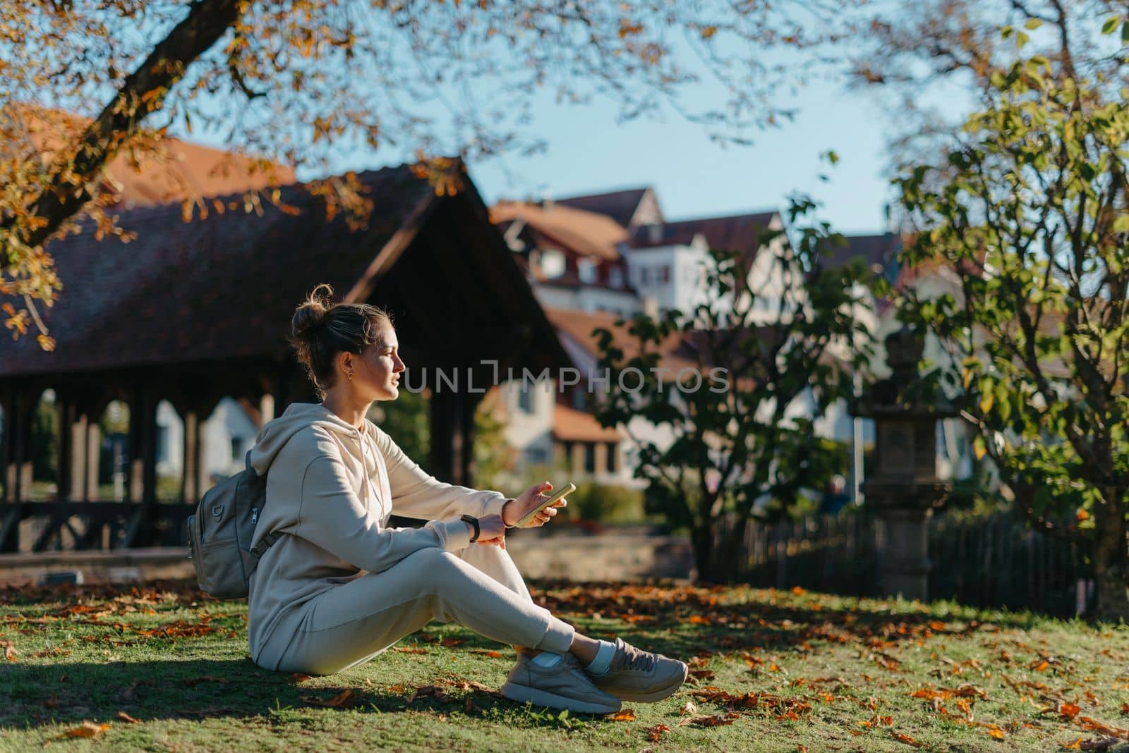 Young fashionable teenage girl with smartphone in park in autumn sitting at smiling. Trendy young woman in fall in park texting. Retouched, vibrant colors. Beautiful blonde teenage girl wearing casual modern autumn outfit sitting in park in autumn. Retouched, vibrant colors, brownish tones.