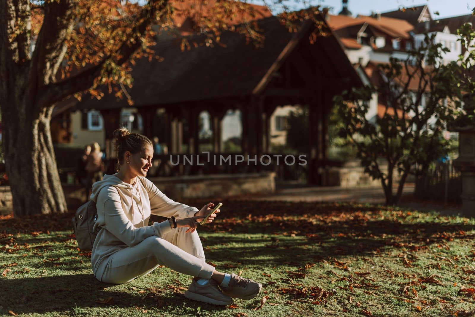 Young Fashionable Teenage Girl With Smartphone In Europian Park In Autumn Sitting At Smiling. Trendy Young Woman In Fall In Park Texting. Retouched, Vibrant Colors. Beautiful Blonde Teenage Girl Wearing Casual Modern Autumn Outfit Sitting In Park In Autumn. Retouched, Vibrant Colors, Brownish Tones. by Andrii_Ko