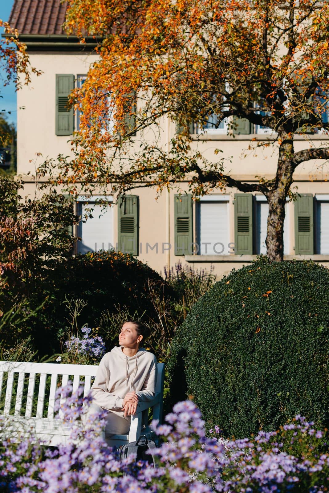 A Girl Sits On A Bench In The Park And Enjoys The Sun. Portrait Young Adult Attractive Woman Enjoy Sitting On Bench And Relaxing Calm Carefree Rest In City Park Against Green Grass And Trees On Sunny Day. Single Female Person Relaxing Chilling Outdoors by Andrii_Ko