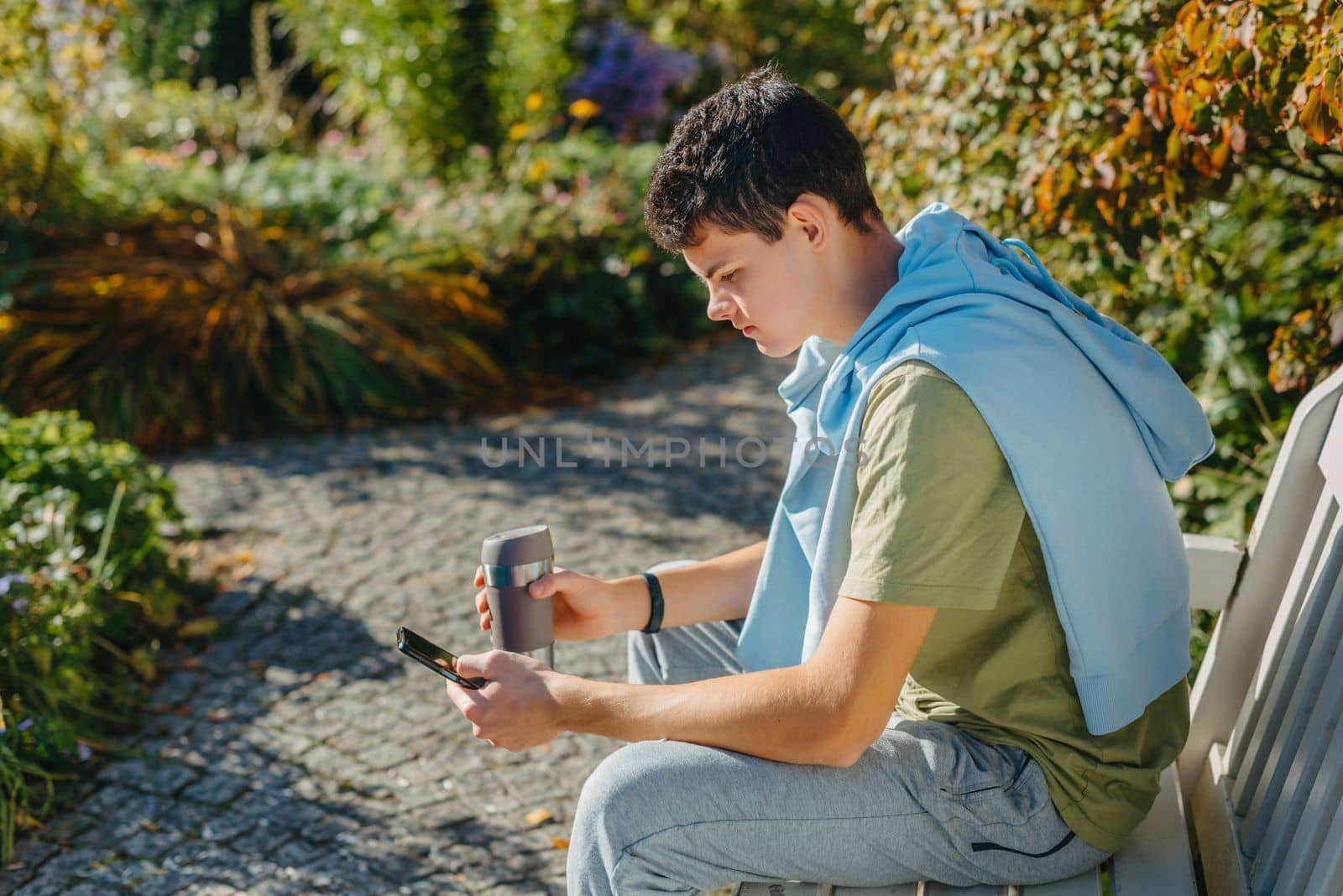 a teenager sits on a bench in the park drinks coffee from a thermo mug and looks into a phone. Portrait of handsome cheerful guy sitting on bench fresh air using device browsing media smm drinking latte urban outside outdoor.