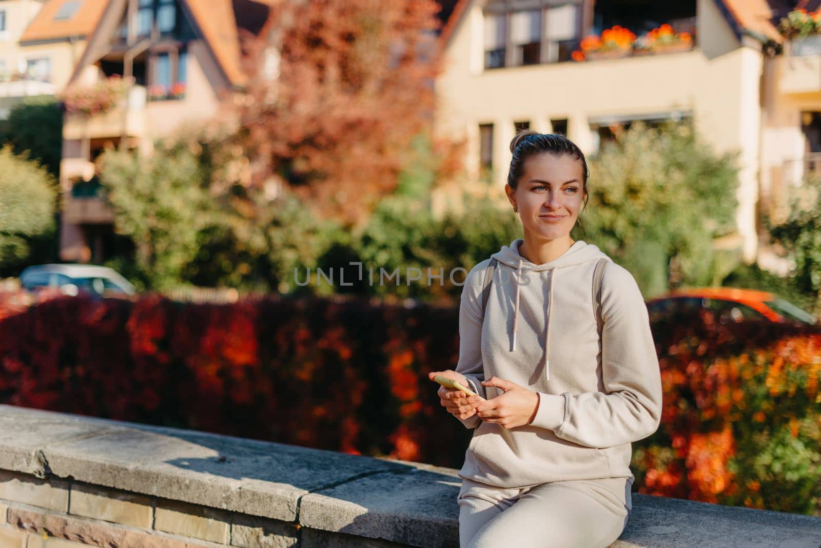 Young fashionable teenage girl with smartphone in park in autumn sitting at smiling. Trendy young woman in fall in park texting. Retouched, vibrant colors. Beautiful blonde teenage girl wearing casual modern autumn outfit sitting in park in autumn. Retouched, vibrant colors, brownish tones.