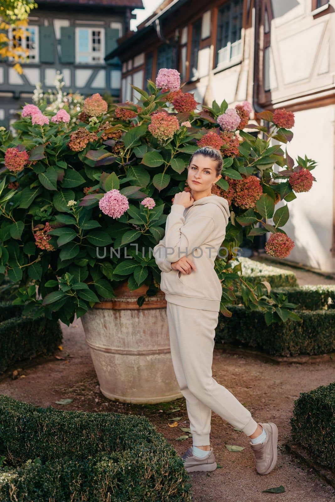 Attractive curly blonde woman walk on the city park street. Girl wear purple hoodie look happy and smiles. Woman make here me gesture standing near pink blooming bush flowers. Happy laughing girl