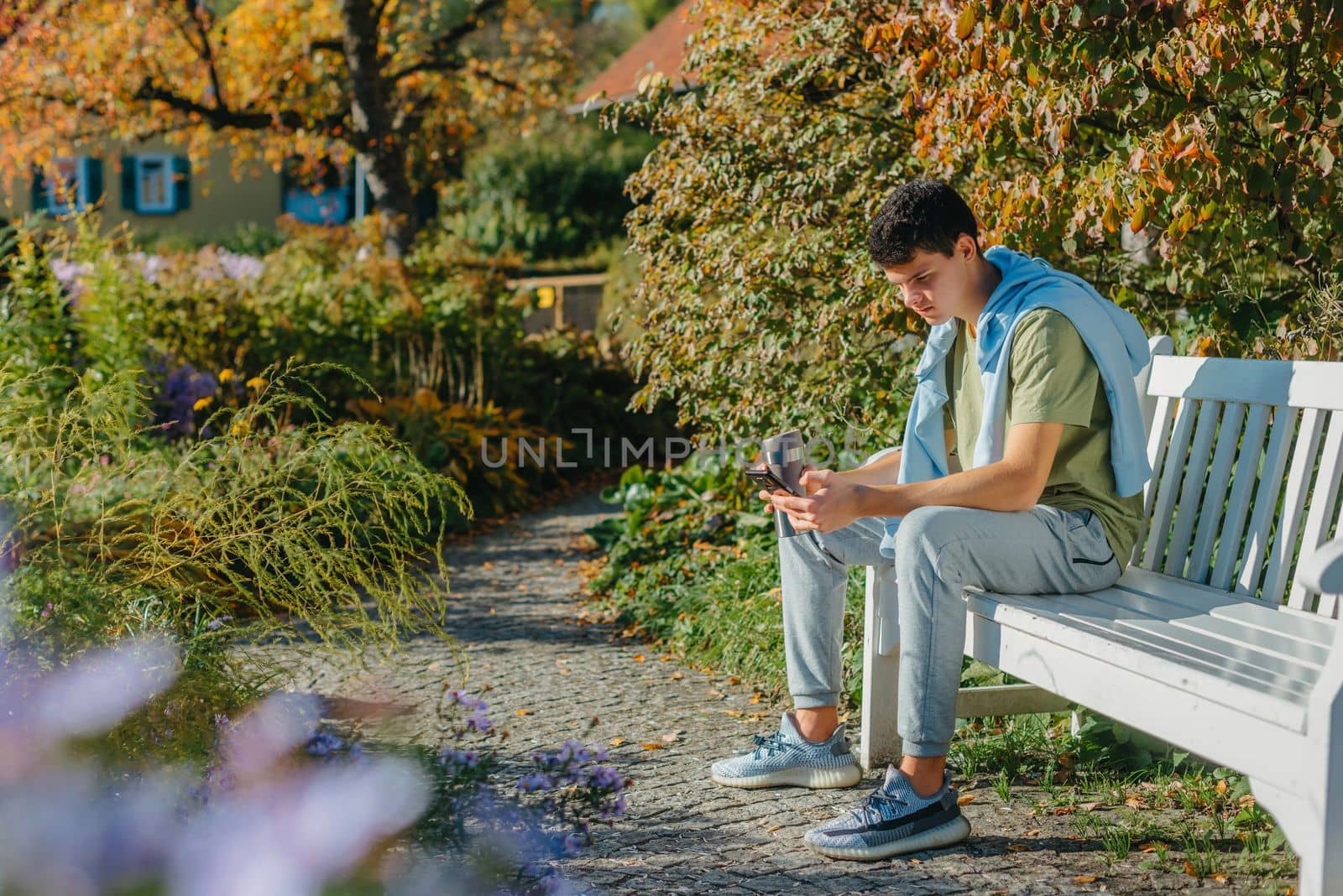 a teenager sits on a bench in the park drinks coffee from a thermo mug and looks into a phone. Portrait of handsome cheerful guy sitting on bench fresh air using device browsing media smm drinking latte urban outside outdoor.