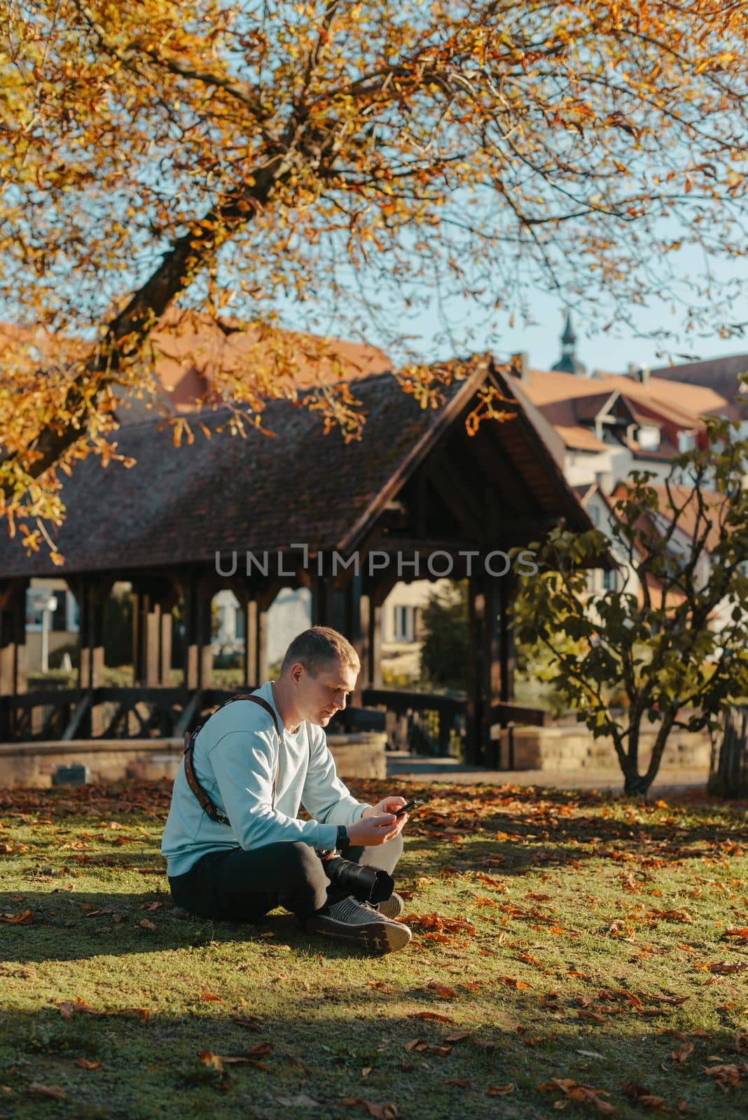 Professional Photographer Taking Picture Of Beautiful Autumn Park. Man Professional Photographer Sit With Camera And With Smartphone In Autumn Park. Retouched, Vibrant Colors, Brownish Tones. by Andrii_Ko