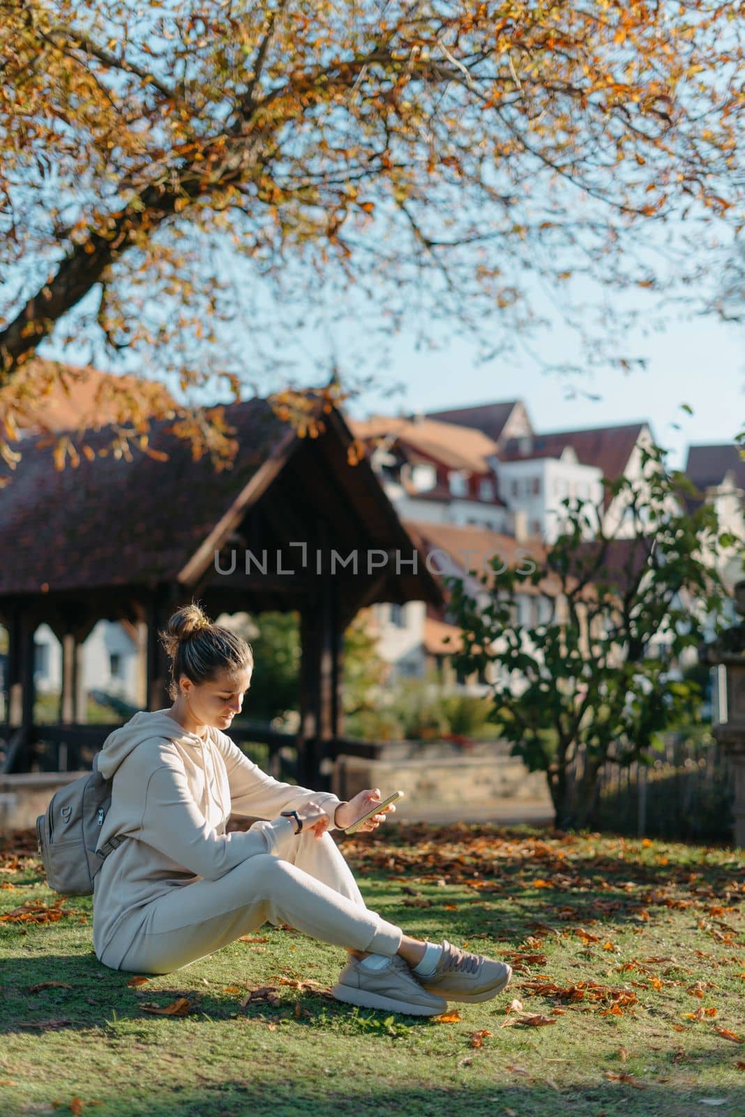 Young Fashionable Teenage Girl With Smartphone In Europian Park In Autumn Sitting At Smiling. Trendy Young Woman In Fall In Park Texting. Retouched, Vibrant Colors. Beautiful Blonde Teenage Girl Wearing Casual Modern Autumn Outfit Sitting In Park In Autumn. Retouched, Vibrant Colors, Brownish Tones. by Andrii_Ko