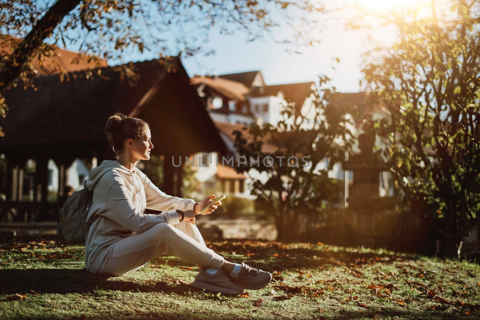 Young fashionable teenage girl with smartphone in park in autumn sitting at smiling. Trendy young woman in fall in park texting. Retouched, vibrant colors. Beautiful blonde teenage girl wearing casual modern autumn outfit sitting in park in autumn. Retouched, vibrant colors, brownish tones.