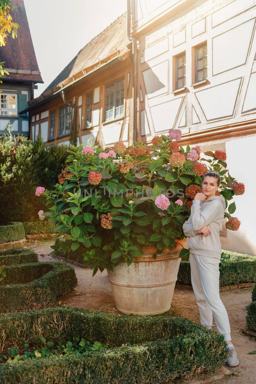 Attractive curly blonde woman walk on the city park street. Girl wear purple hoodie look happy and smiles. Woman make here me gesture standing near pink blooming bush flowers. Happy laughing girl. by Andrii_Ko