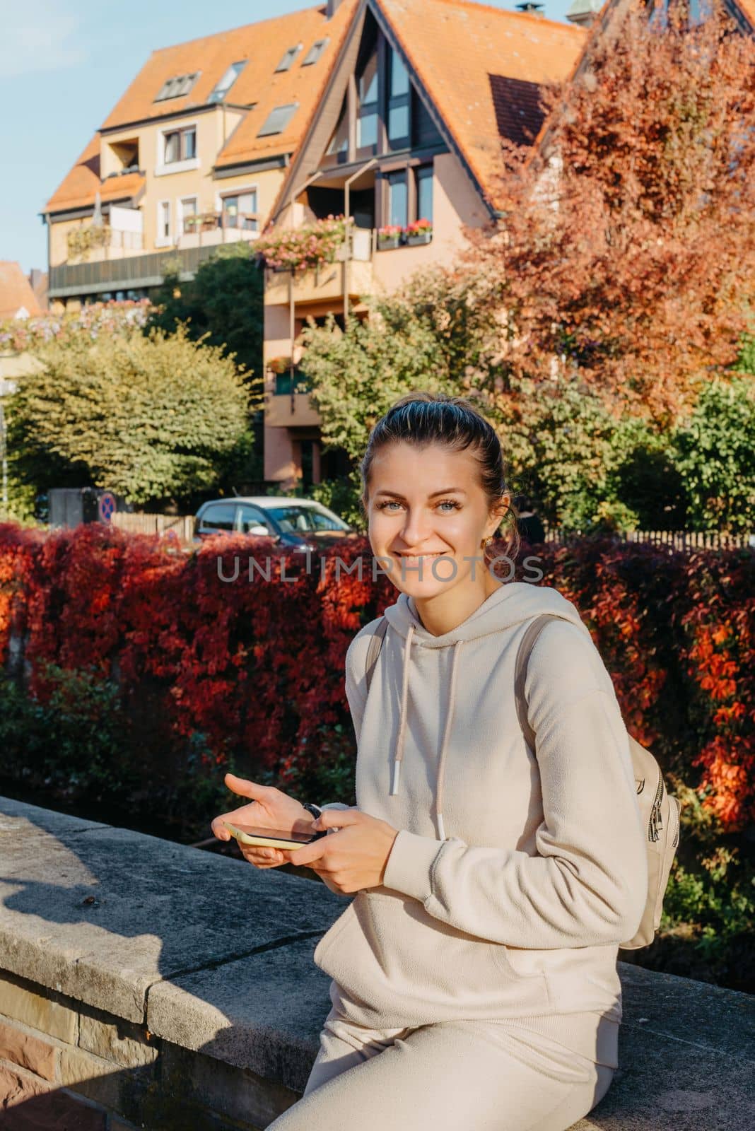 Young fashionable teenage girl with smartphone in park in autumn sitting at smiling. Trendy young woman in fall in park texting. Retouched, vibrant colors. Beautiful blonde teenage girl wearing casual modern autumn outfit sitting in park in autumn. Retouched, vibrant colors, brownish tones.