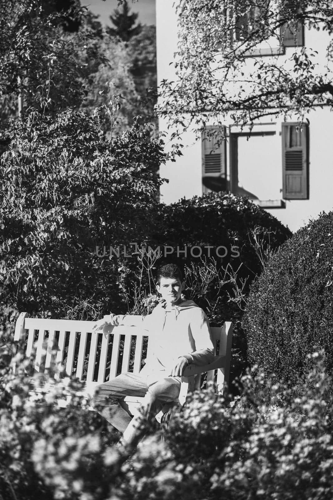 a teenager sits on a bench in the park drinks coffee from a thermo mug and looks into a phone. Portrait of handsome cheerful guy sitting on bench fresh air using device browsing media smm drinking latte urban outside outdoor.