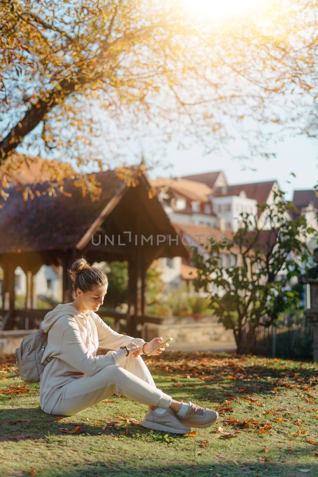 Young Fashionable Teenage Girl With Smartphone In Europian Park In Autumn Sitting At Smiling. Trendy Young Woman In Fall In Park Texting. Retouched, Vibrant Colors. Beautiful Blonde Teenage Girl Wearing Casual Modern Autumn Outfit Sitting In Park In Autumn. Retouched, Vibrant Colors, Brownish Tones. by Andrii_Ko