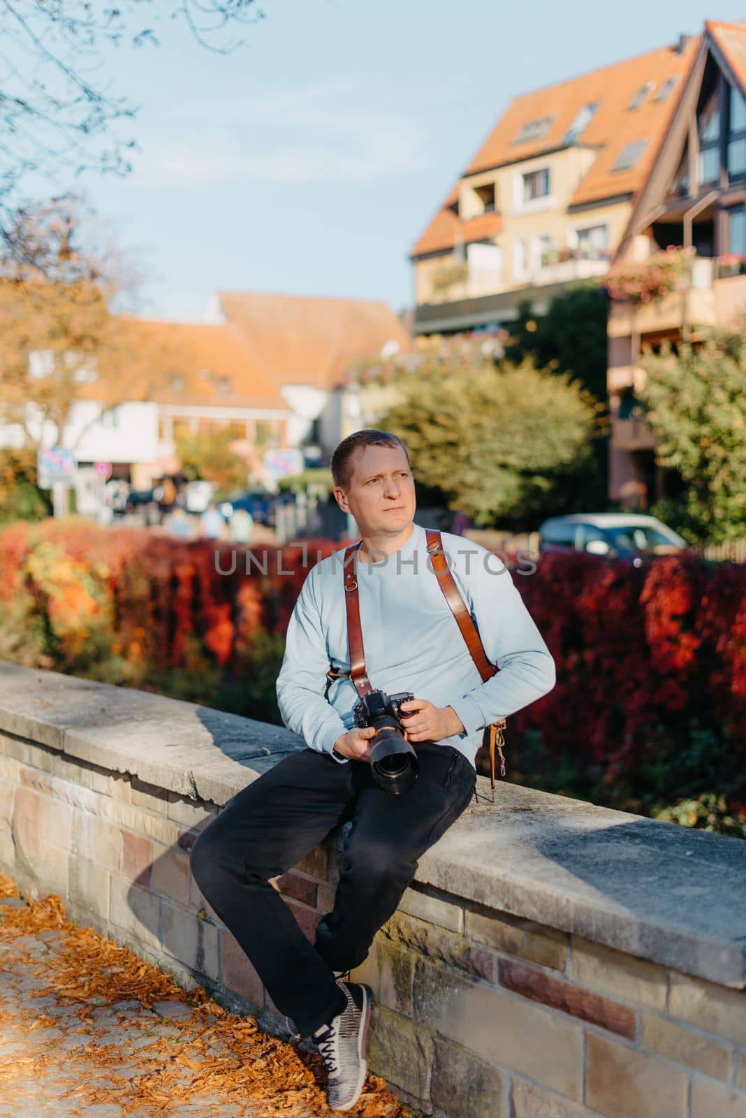 Man Sitting in Old European City And Holding Photo Camera. Contemporary Stylish Blogger And Photographer. Handsome man taking a selfie on a trip in Europe. by Andrii_Ko