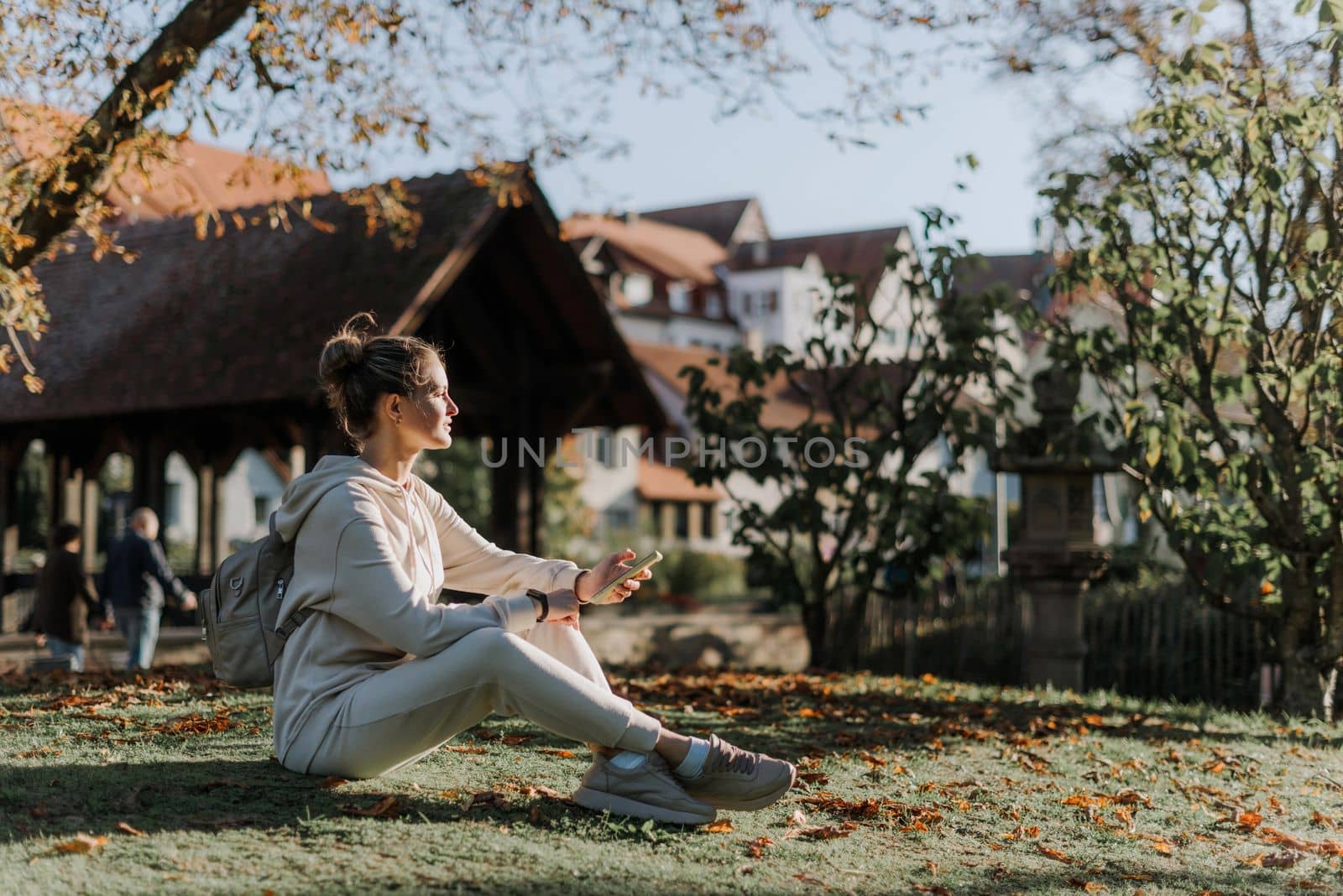 Young fashionable teenage girl with smartphone in park in autumn sitting at smiling. Trendy young woman in fall in park texting. Retouched, vibrant colors. Beautiful blonde teenage girl wearing casual modern autumn outfit sitting in park in autumn. Retouched, vibrant colors, brownish tones.