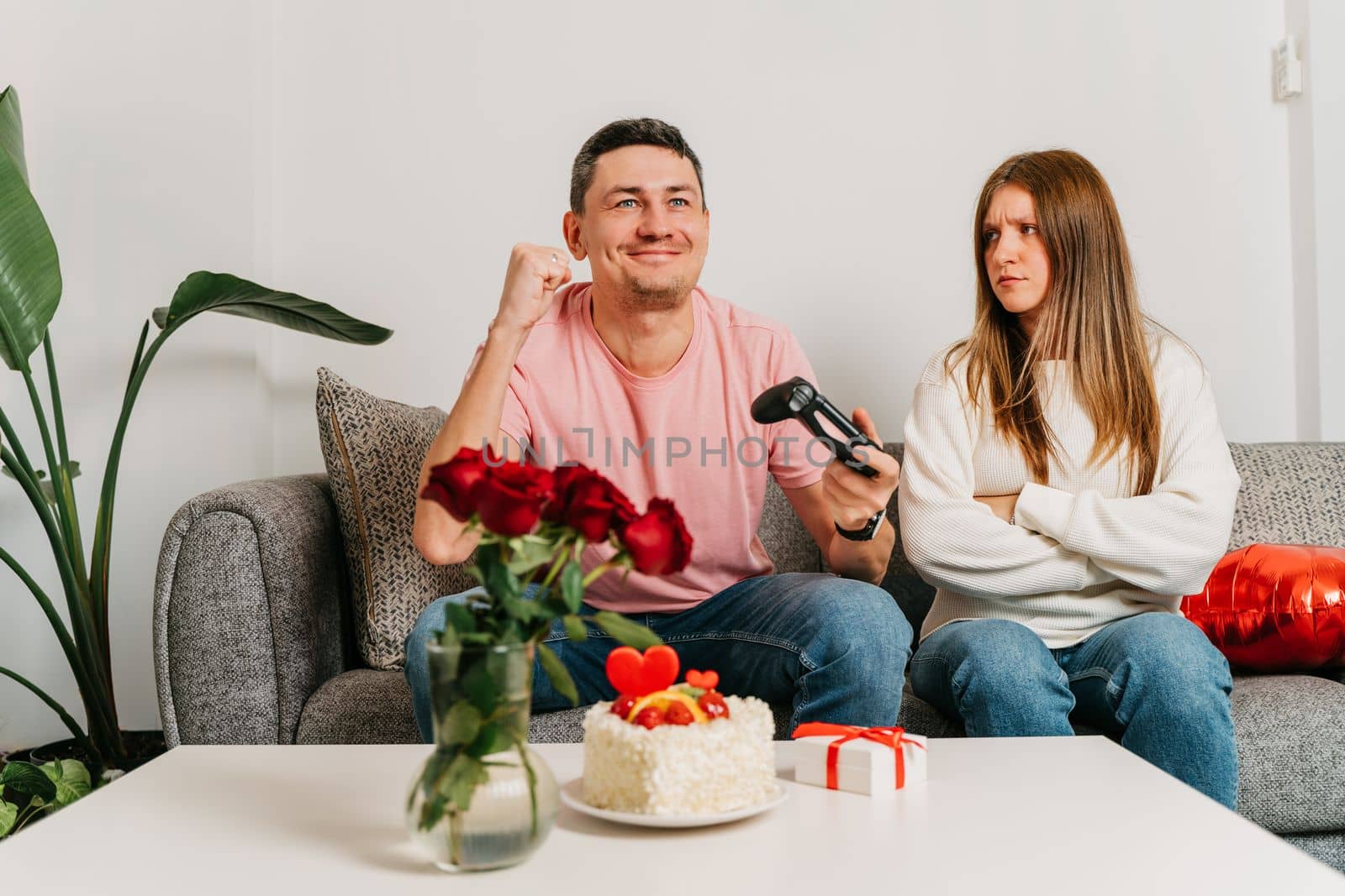 Beautiful young couple sitting on a couch, having a quarrel over romantic date on Valentine day. Man playing video games at gamepad, woman in anger.