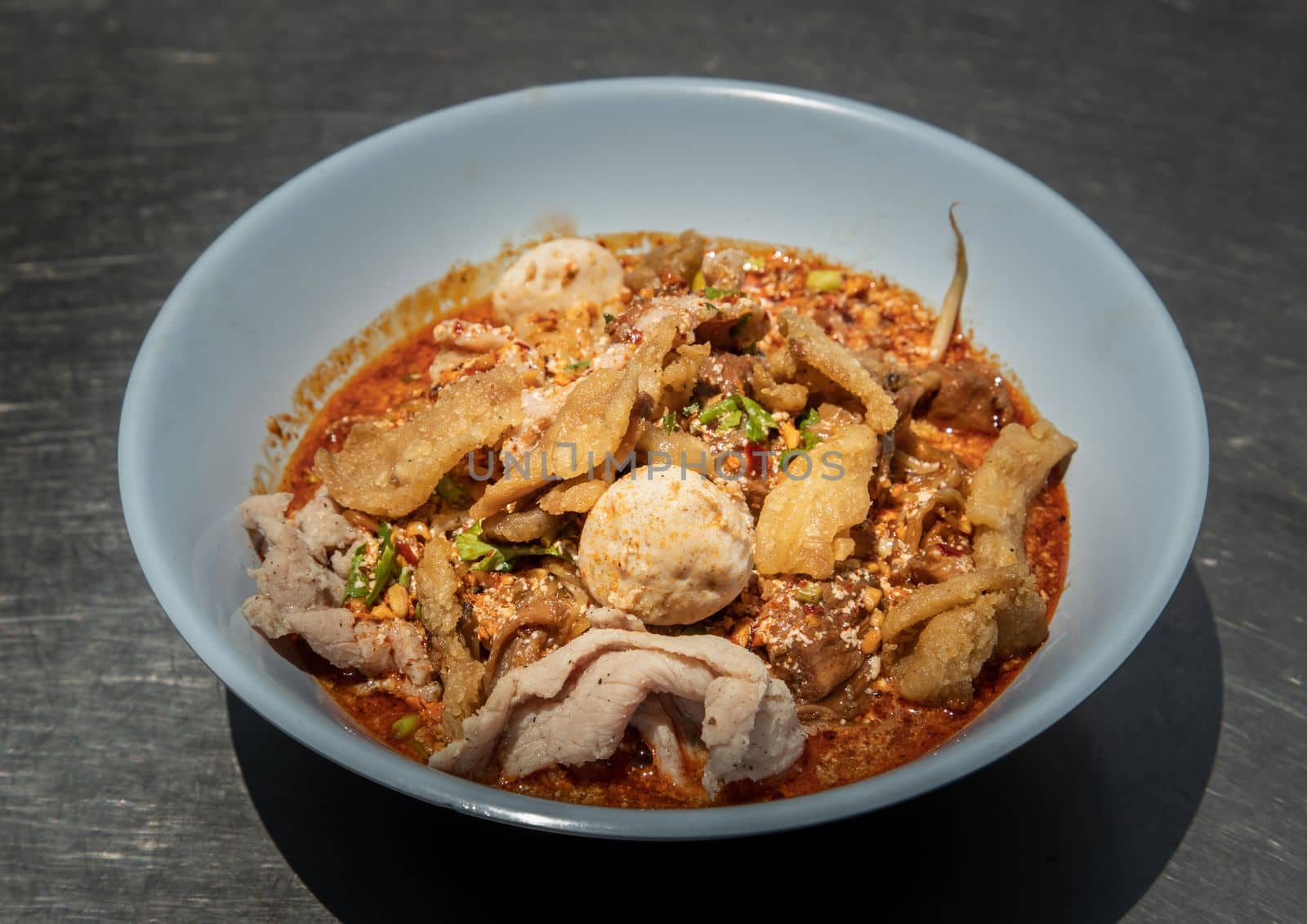 Egg noodles in Thai spicy soup with braised pork, pork balls and fatty pork rinds in white bowl. Thai food, Selective focus.