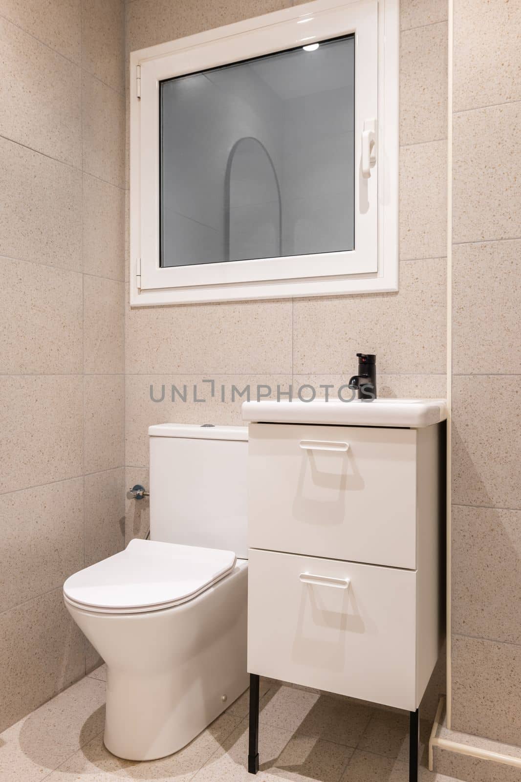 Part of bathroom with toilet bowl, washbasin on wooden furniture and beige tiled walls.