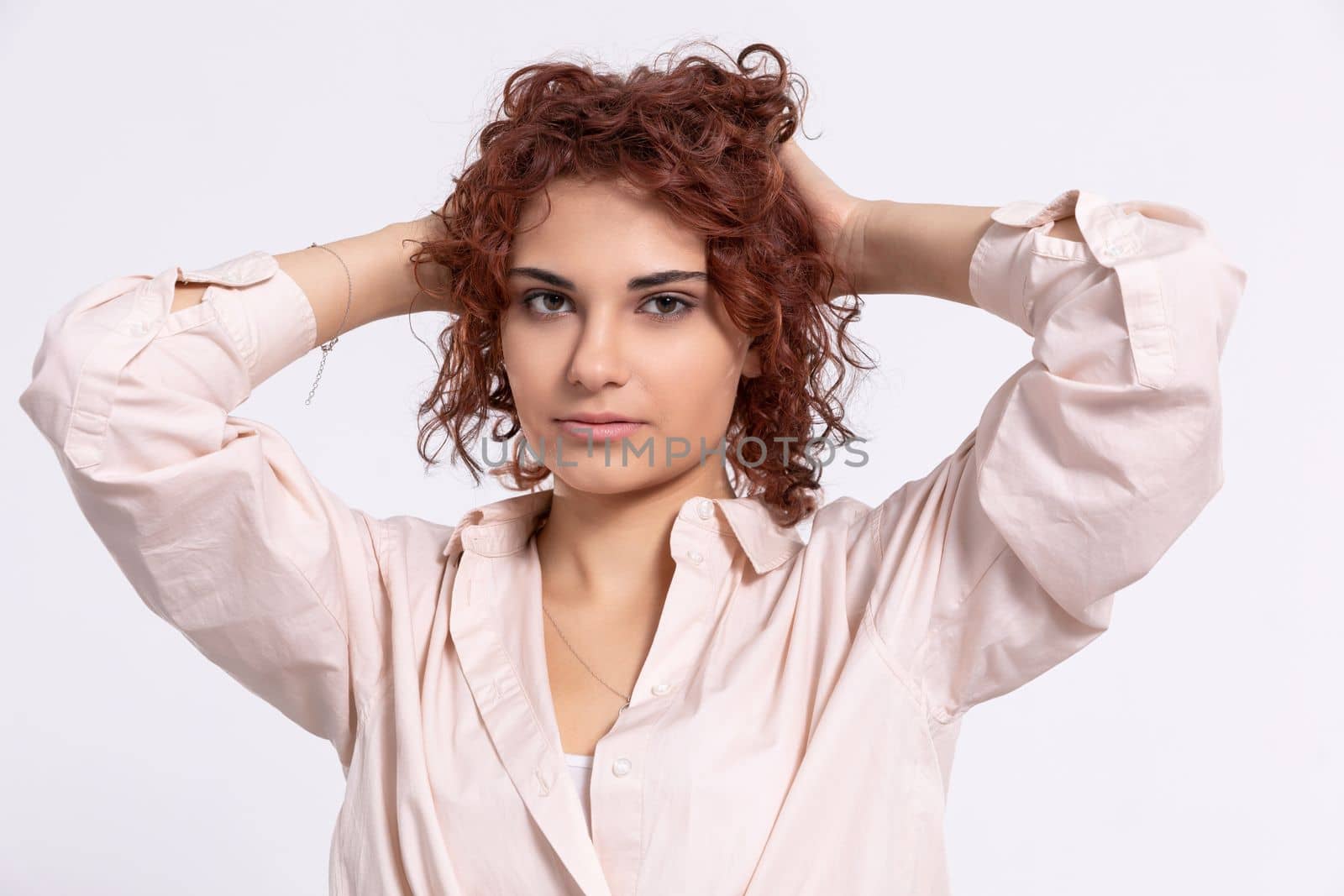 The girl raises her hair up with her hands. Beautiful short curly chestnut-colored hair. Natural hairstyle. Light-colored long-sleeved blouse.