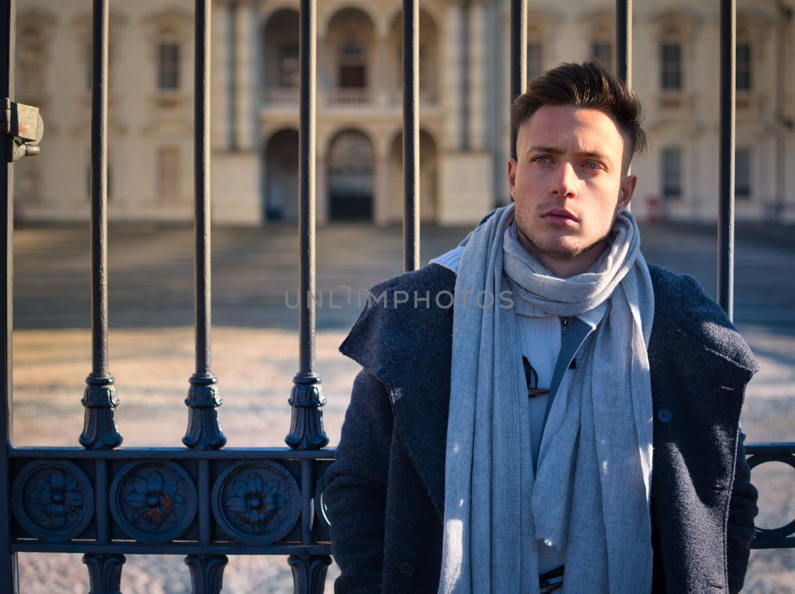 One handsome young man in urban setting in European city, standing, wearing black coat and scarf in winter day, leaning against metal gate