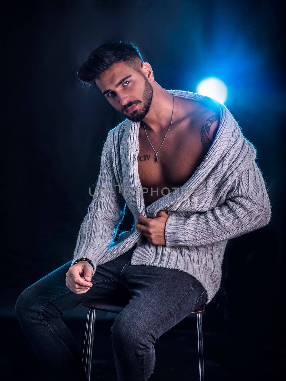 Attractive young man sitting on stool, wearing wool sweater open on naked chest, on black background, looking at camera