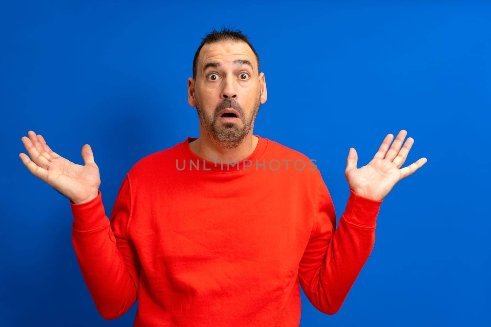 Amazed and excited latin man looking at camera with a shocked expression feeling shocked from advertising shopping promotion, amazing betting win standing isolated on a blue background.