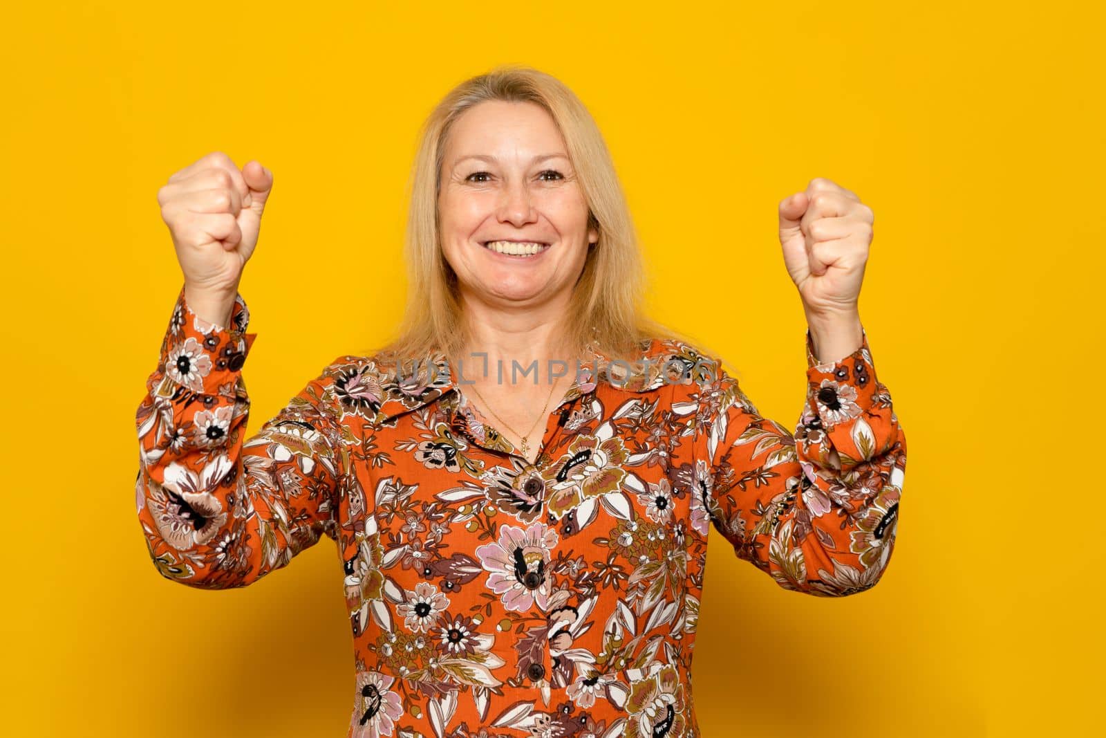 Happy caucasian successful woman in a patterned dress with raised hands smiling and celebrating success over yellow background