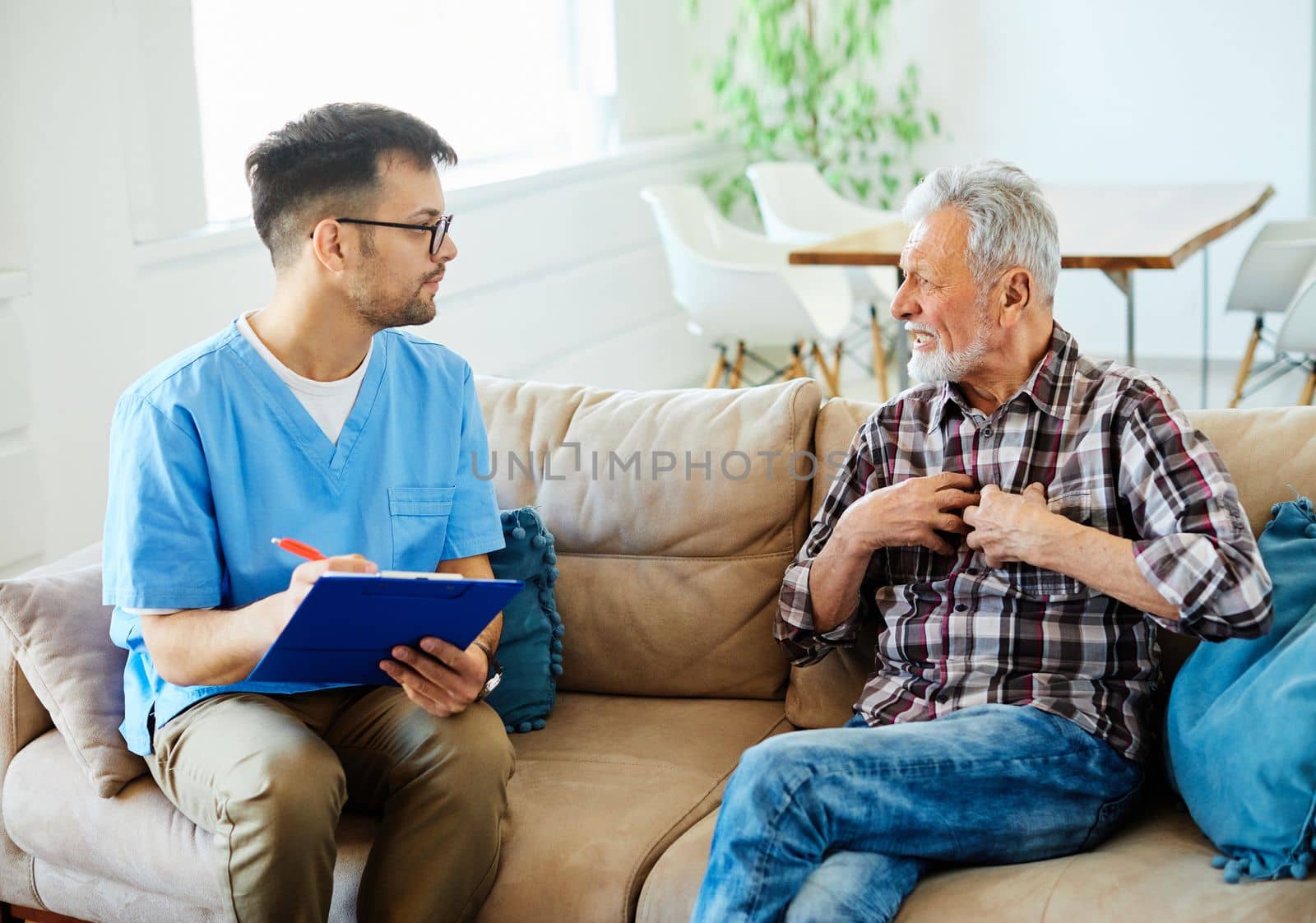 Doctor or nurse caregiver talking with senior man with chest pain and coughing problems on sofa at home or nursing home