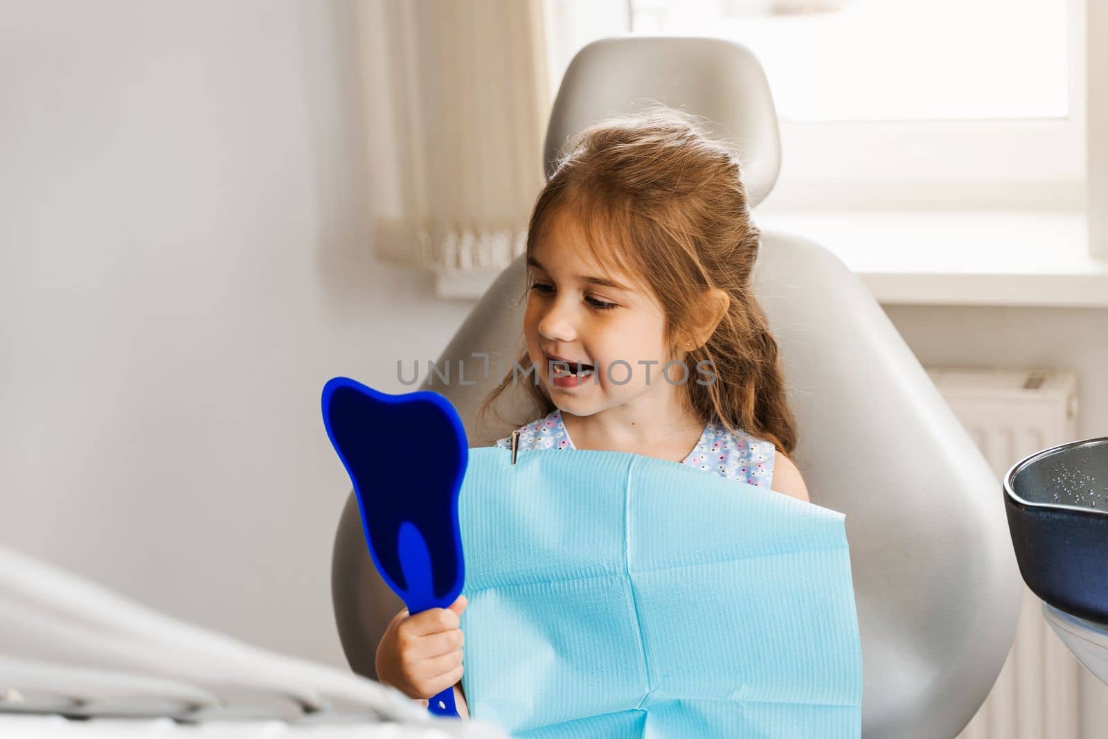 Teeth treatment. Child looking in the mirror at the dentist. Happy child patient of dentistry. Attractive kid girl sitting in dental office and smiling