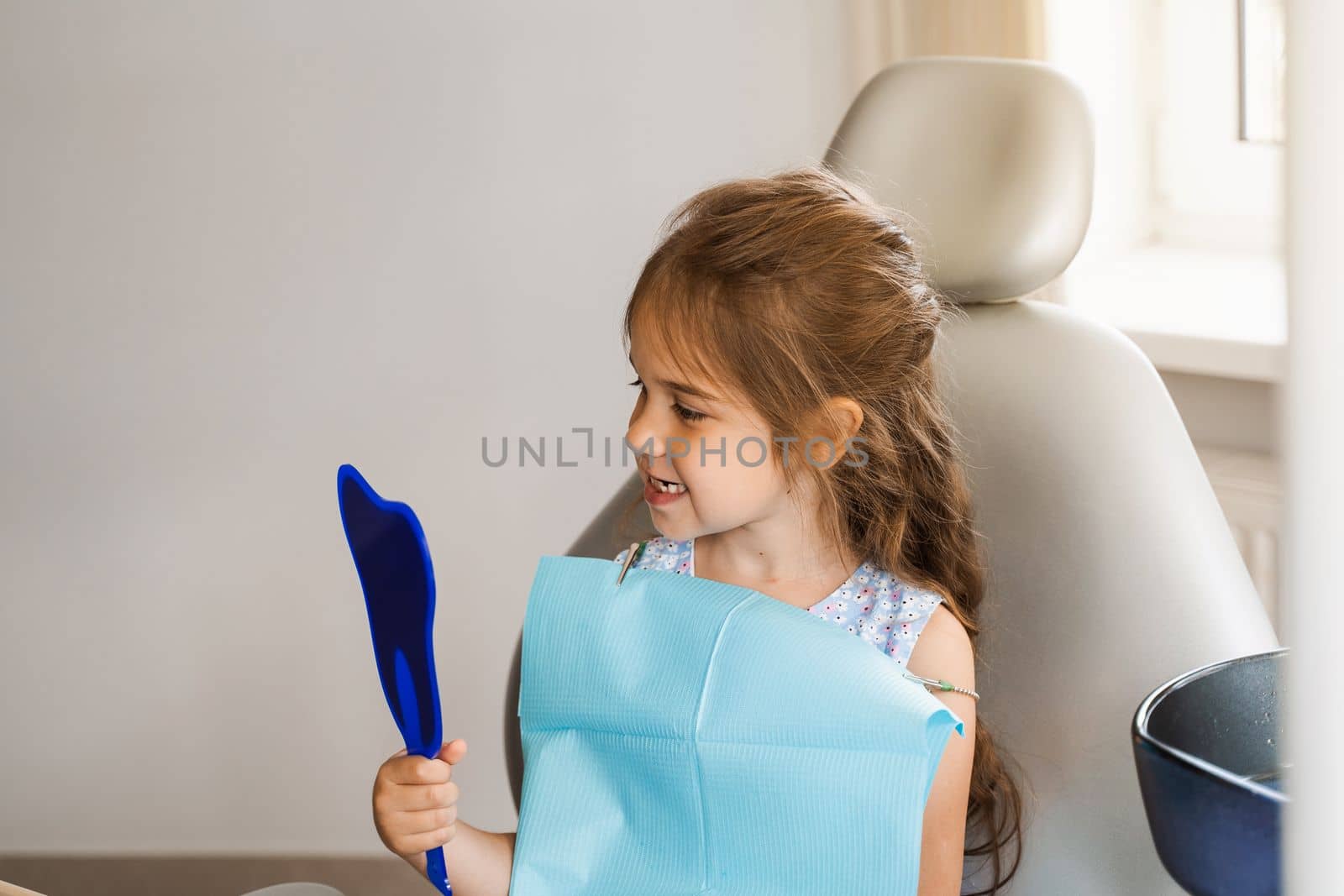 Child looking in the mirror at the dentist. Happy child patient of dentistry. Teeth treatment. Attractive kid girl sitting in dental office and smiling. by Rabizo