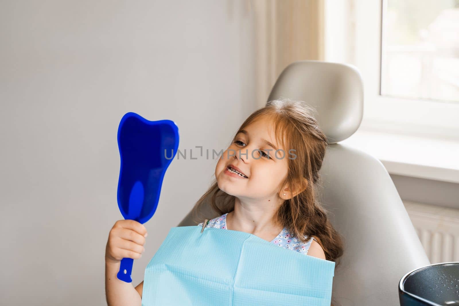 Teeth treatment. Child looking in the mirror at the dentist. Happy child patient of dentistry. Attractive kid girl sitting in dental office and smiling. by Rabizo