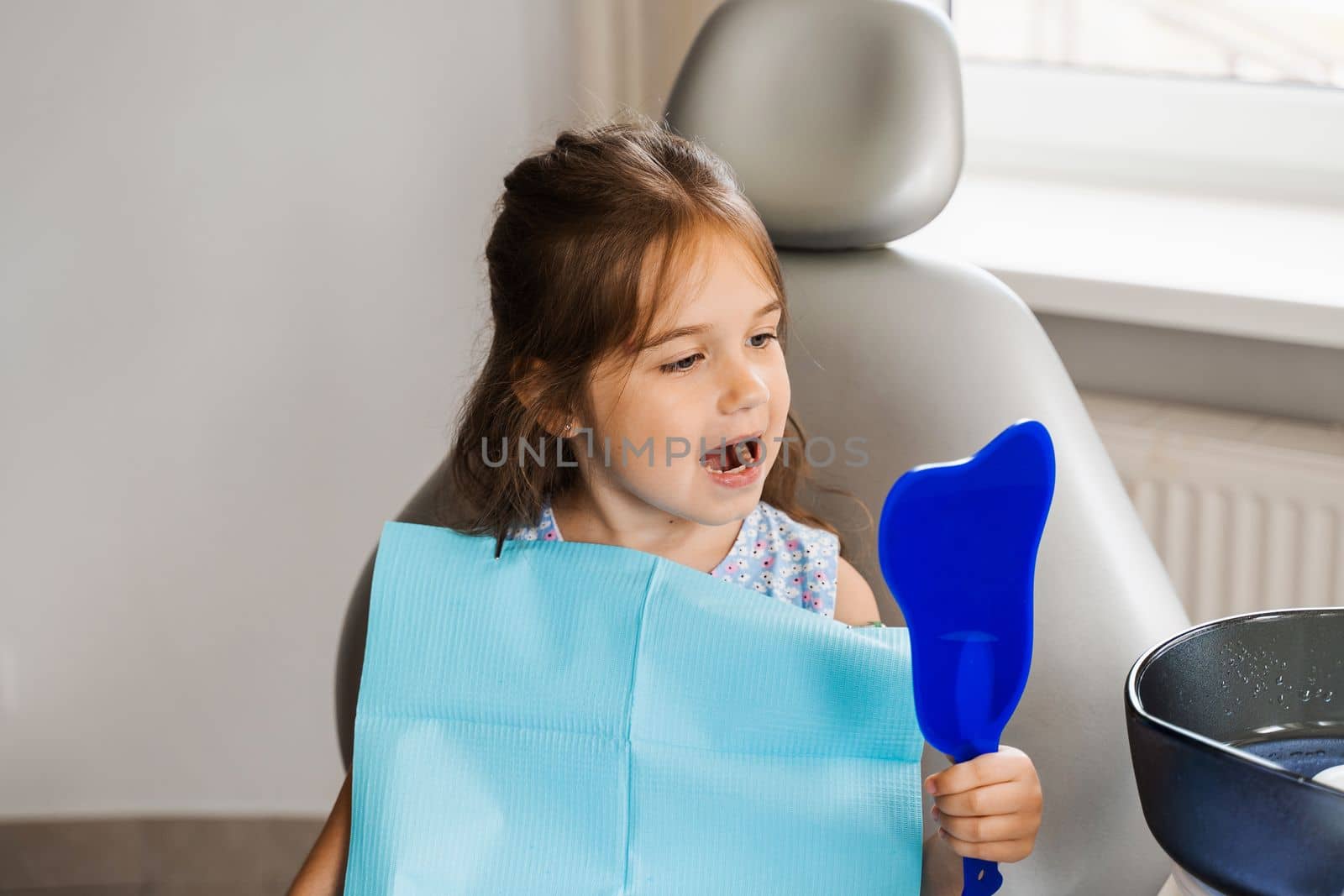 Teeth treatment. Child looking in the mirror at the dentist. Happy child patient of dentistry. Attractive kid girl sitting in dental office and smiling. by Rabizo
