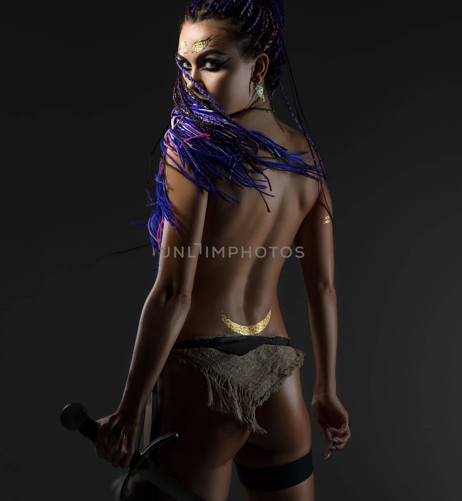 Sexy resolute woman with dreadlocks topless holding a sword in her hand in the image of amazon rearview on gray background