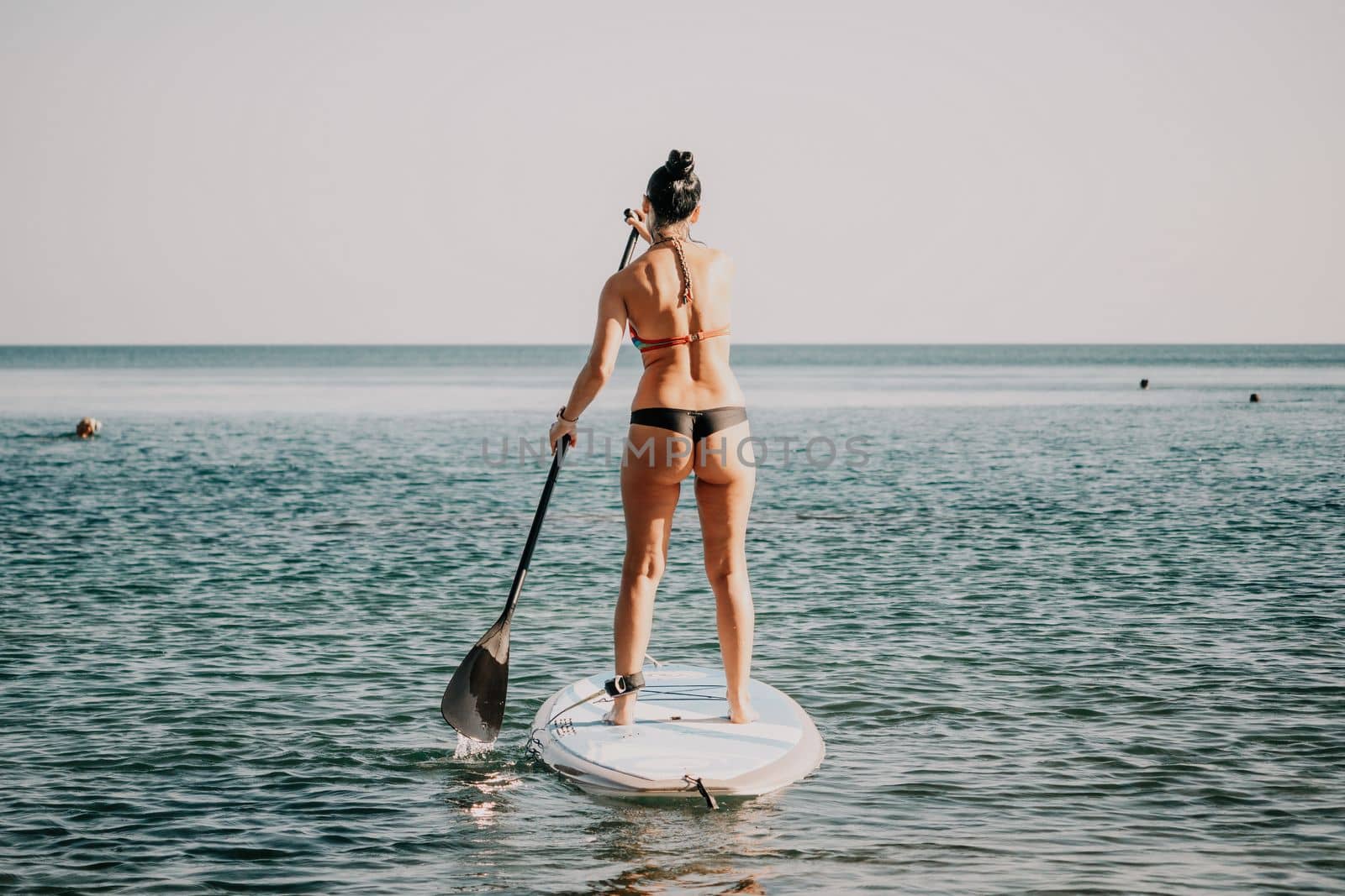 Sea woman sup. Silhouette of happy middle aged woman in rainbow bikini, surfing on SUP board, confident paddling through water surface. Idyllic sunset. Active lifestyle at sea or river