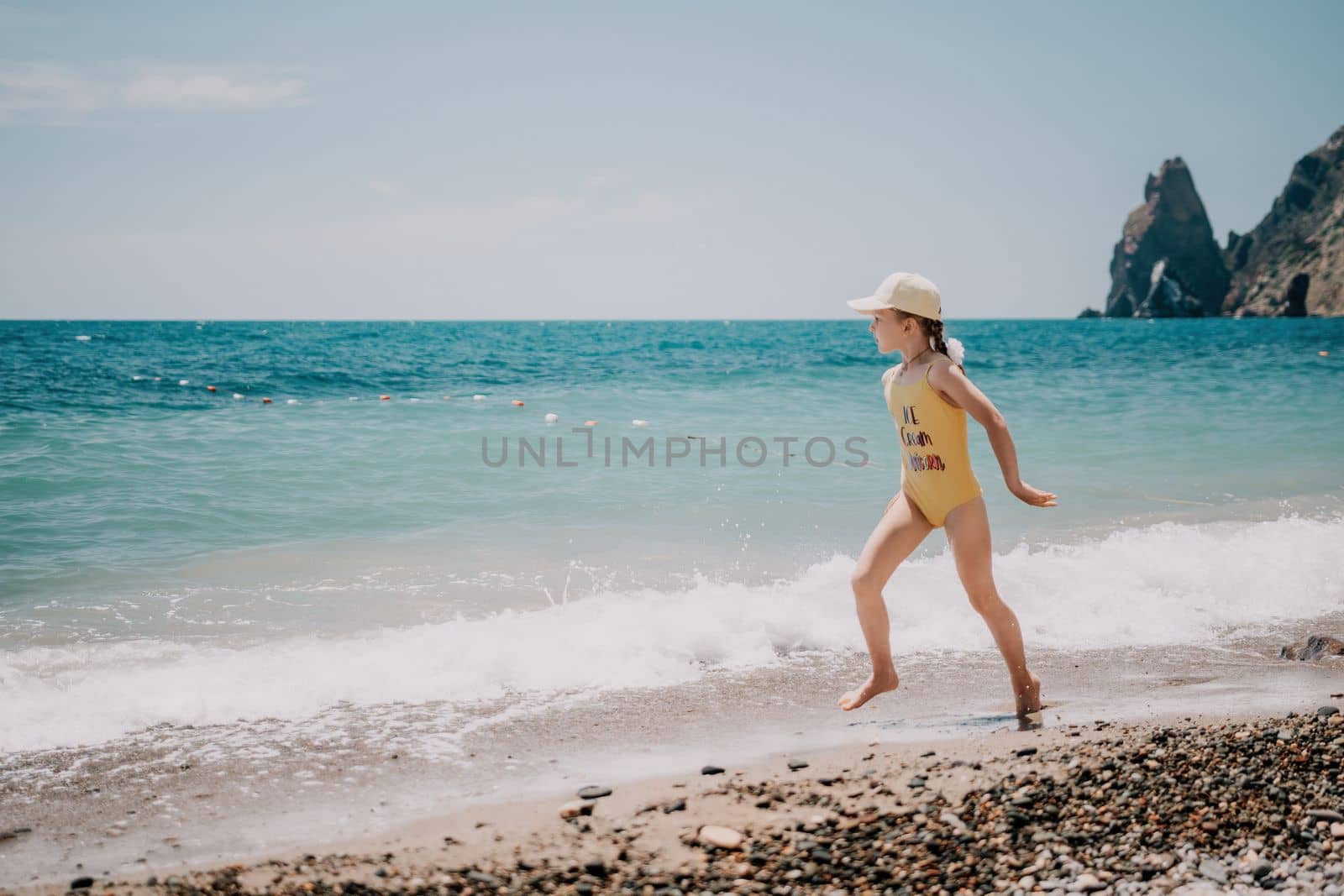 Cute little girl running along the seashore against a clear blue sea and rejoices in the rays of the summer sun. Beautiful girl in yellow swimsuit running and having fun on tropical beach