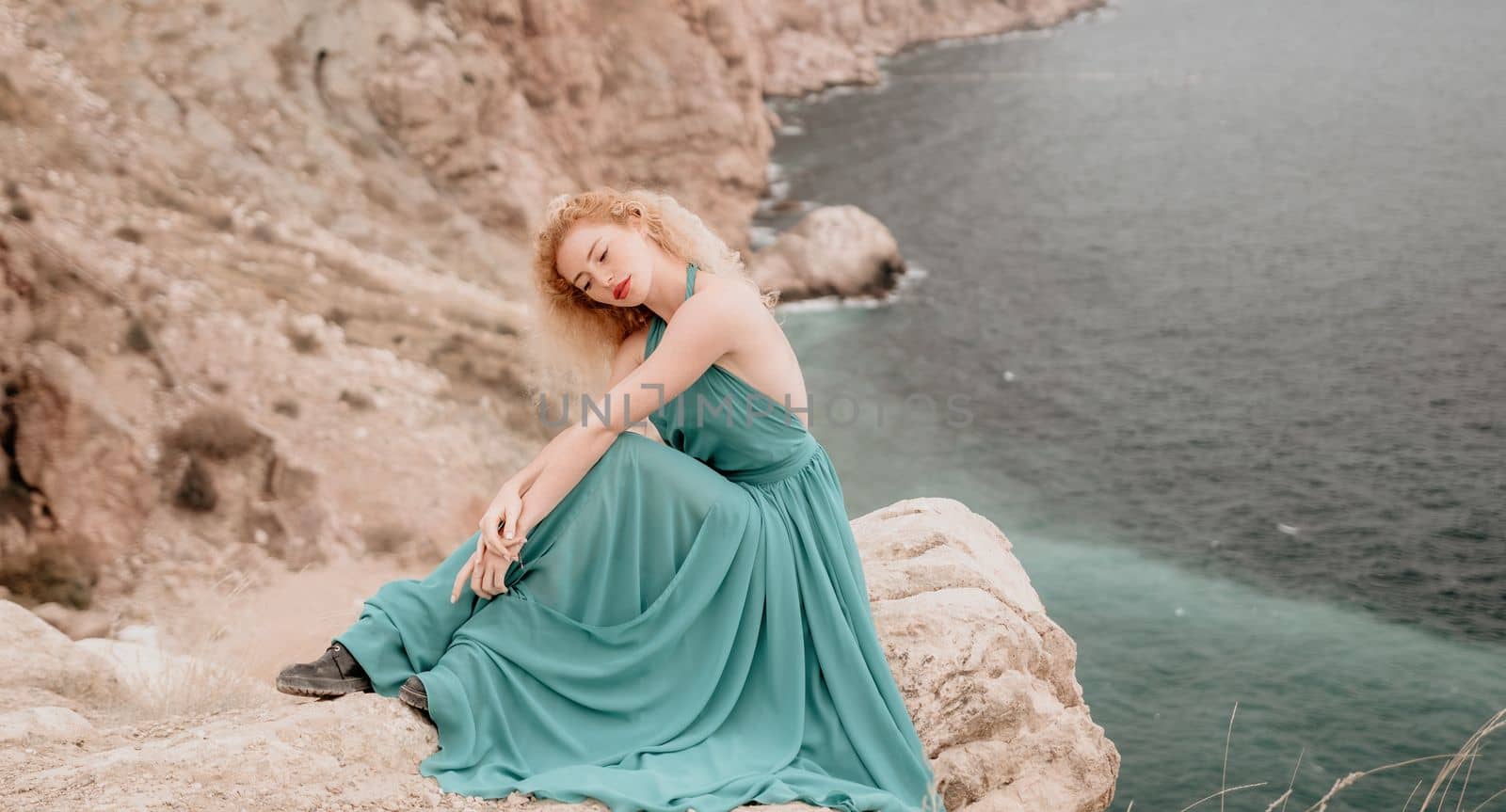 Redhead woman portrait. Curly redhead young caucasian woman with freckles looking at camera and smiling. Close up portrait cute woman in a mint long dress posing on a volcanic rock high above the sea by panophotograph