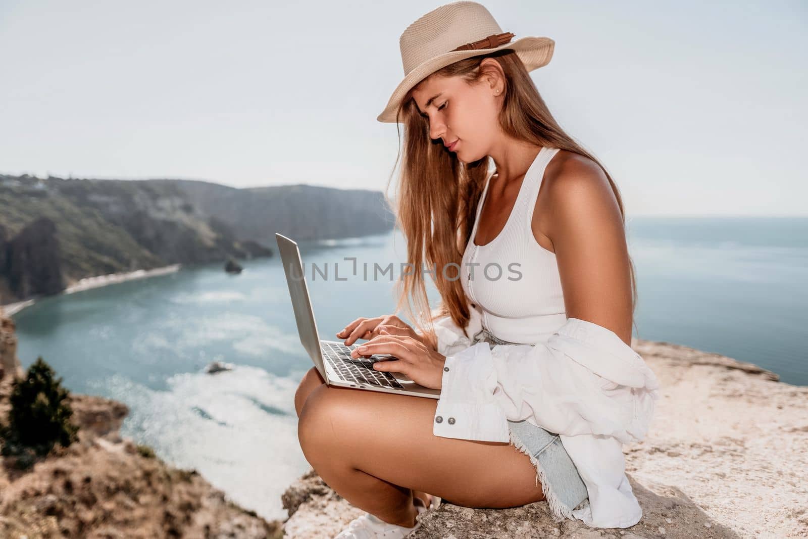 Woman sea laptop. Business woman in yellow hat freelancer with laptop working over blue sea beach. Girl relieves stress from work. Freelance, digital nomad, travel and holidays concept by panophotograph