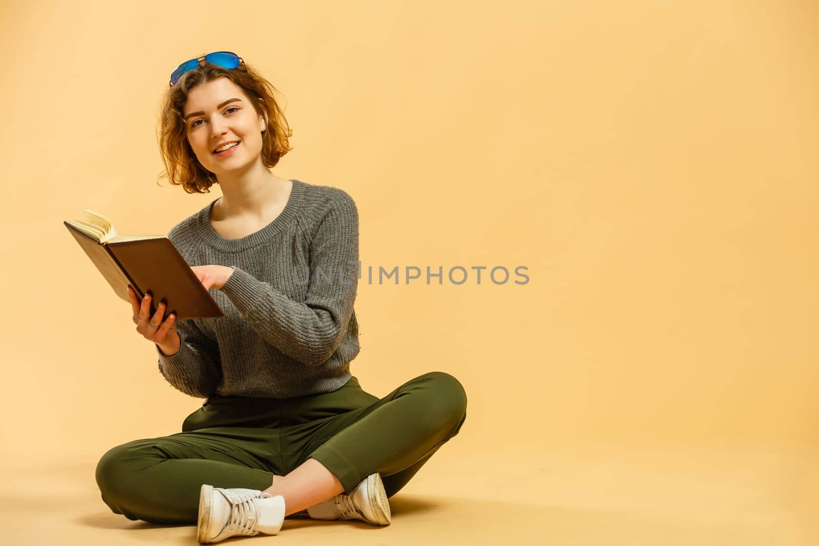 Portrait of cheerful young woman college student holding book and looking at camera on isolated beige background. Pretty redhead lady model wearing casual clothes emotionally showing facial expressions.