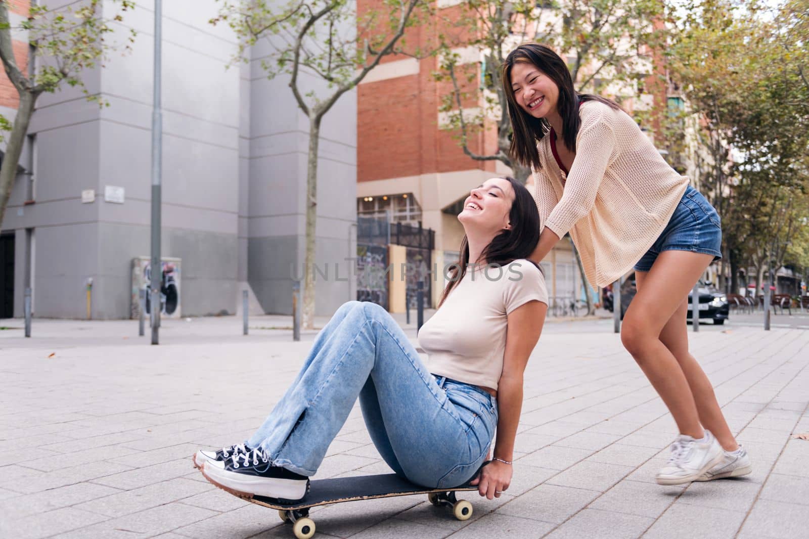 two young women laughing and having fun playing with a skateboard in the street, concept of friendship and teenager lifestyle, copy space for text