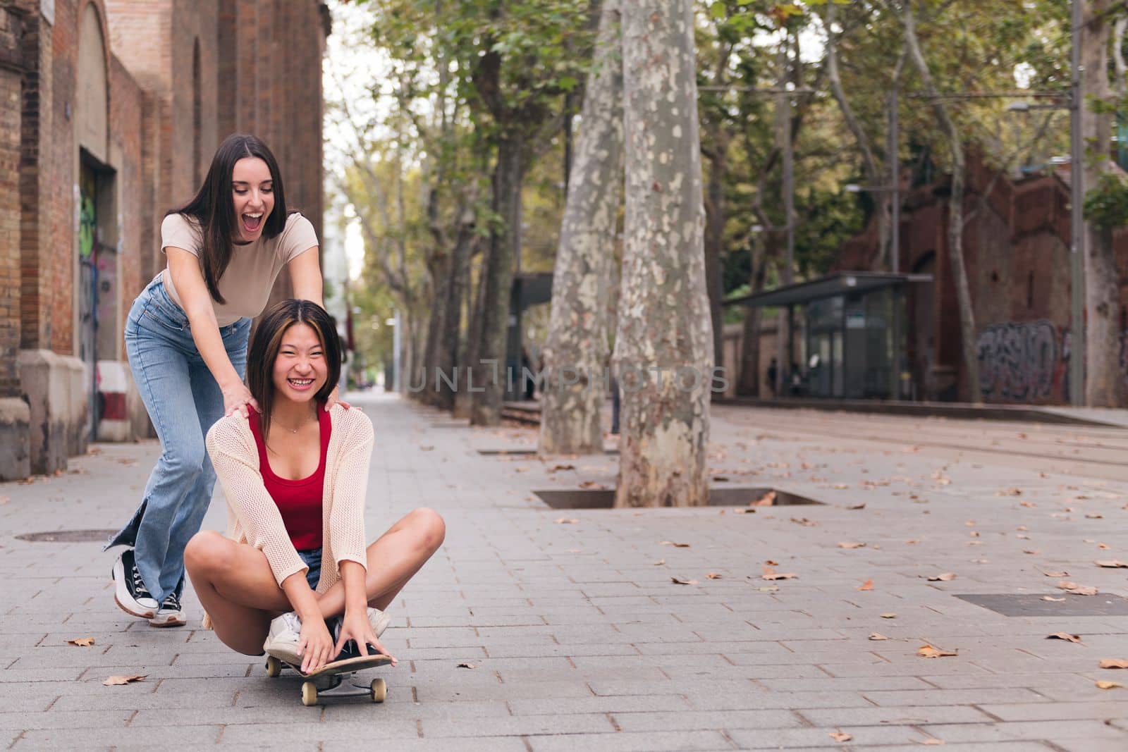young woman laughing and having a good time pushing her friend sitting on a skateboard down a city street, concept of friendship and teenager lifestyle, copy space for text