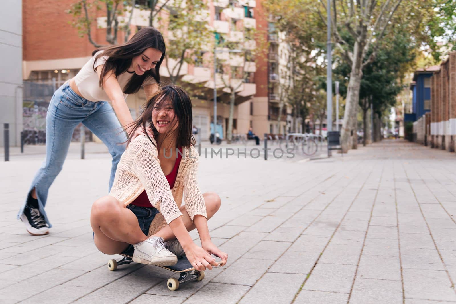 two young women laughing and having a good time playing with a skateboard down a city street, concept of friendship and teenager lifestyle, copy space for text