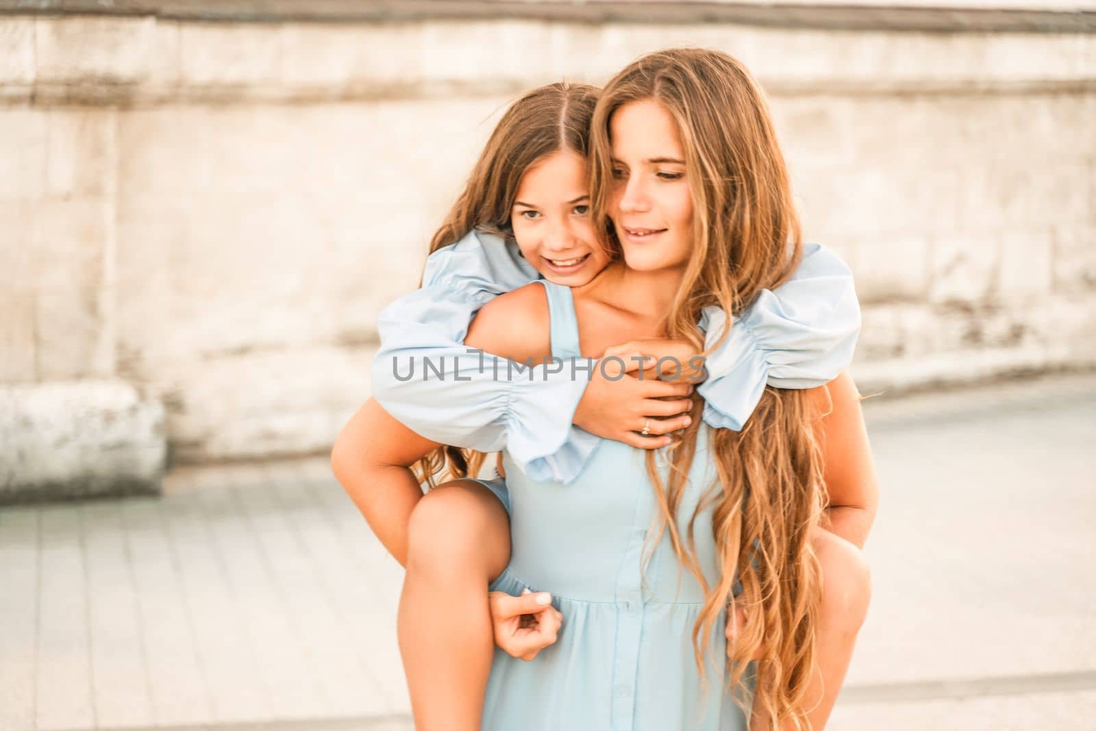 Mother of the daughter walks playing. Mother holds the girl on her back, holding her legs, and her daughter hugs her by the shoulders. Dressed in blue dresses