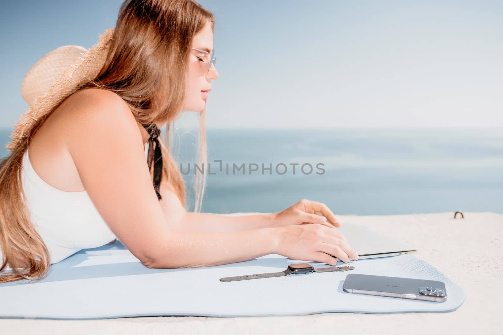 Successful business woman in yellow hat working on laptop by the sea. Pretty lady typing on computer at summer day outdoors. Freelance, travel and holidays concept.