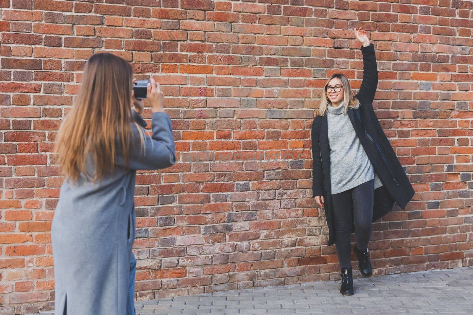 Girl takes picture of her female friend in front of brick wall in urban street - photographer and youth urban lifestyle