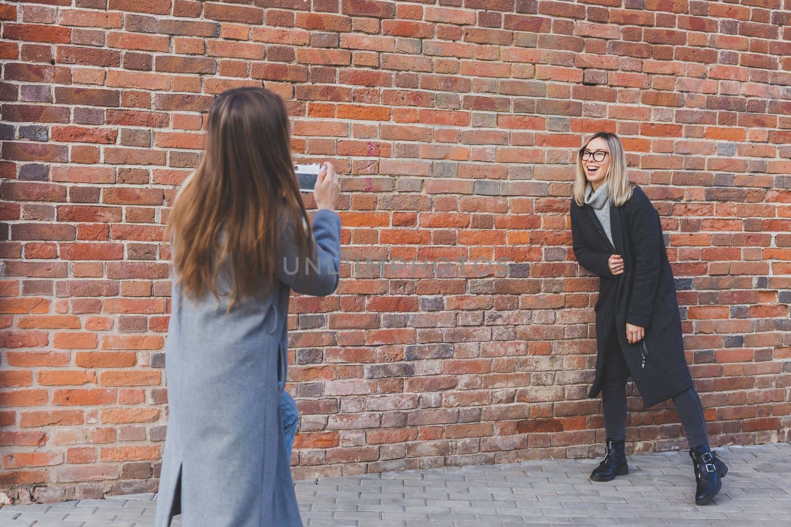 Girl takes picture of her female friend in front of brick wall in city street - photographer and vintage camera hobby concept by Satura86