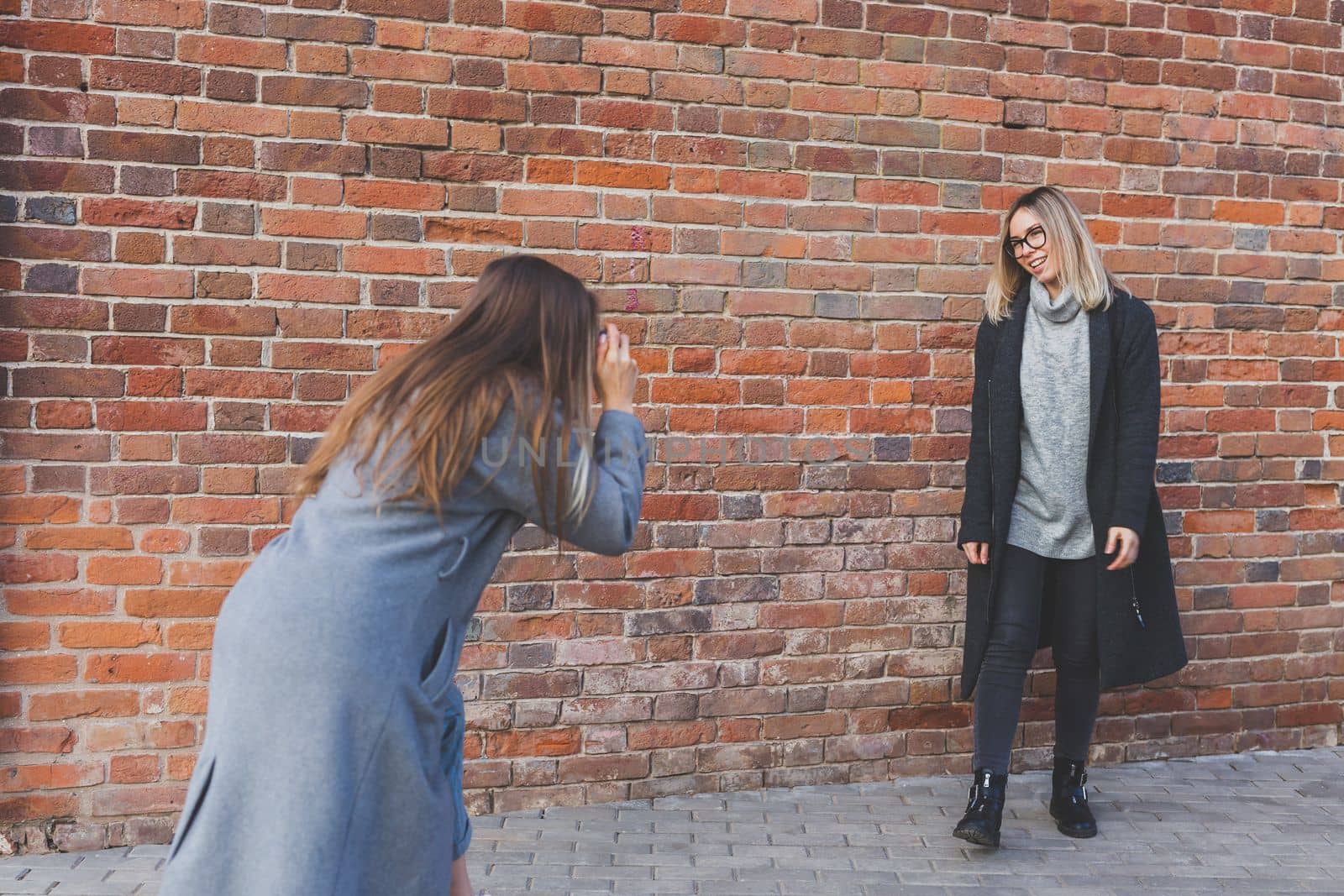 Girl takes picture of her female friend in front of brick wall in city street - photographer and vintage camera hobby concept by Satura86