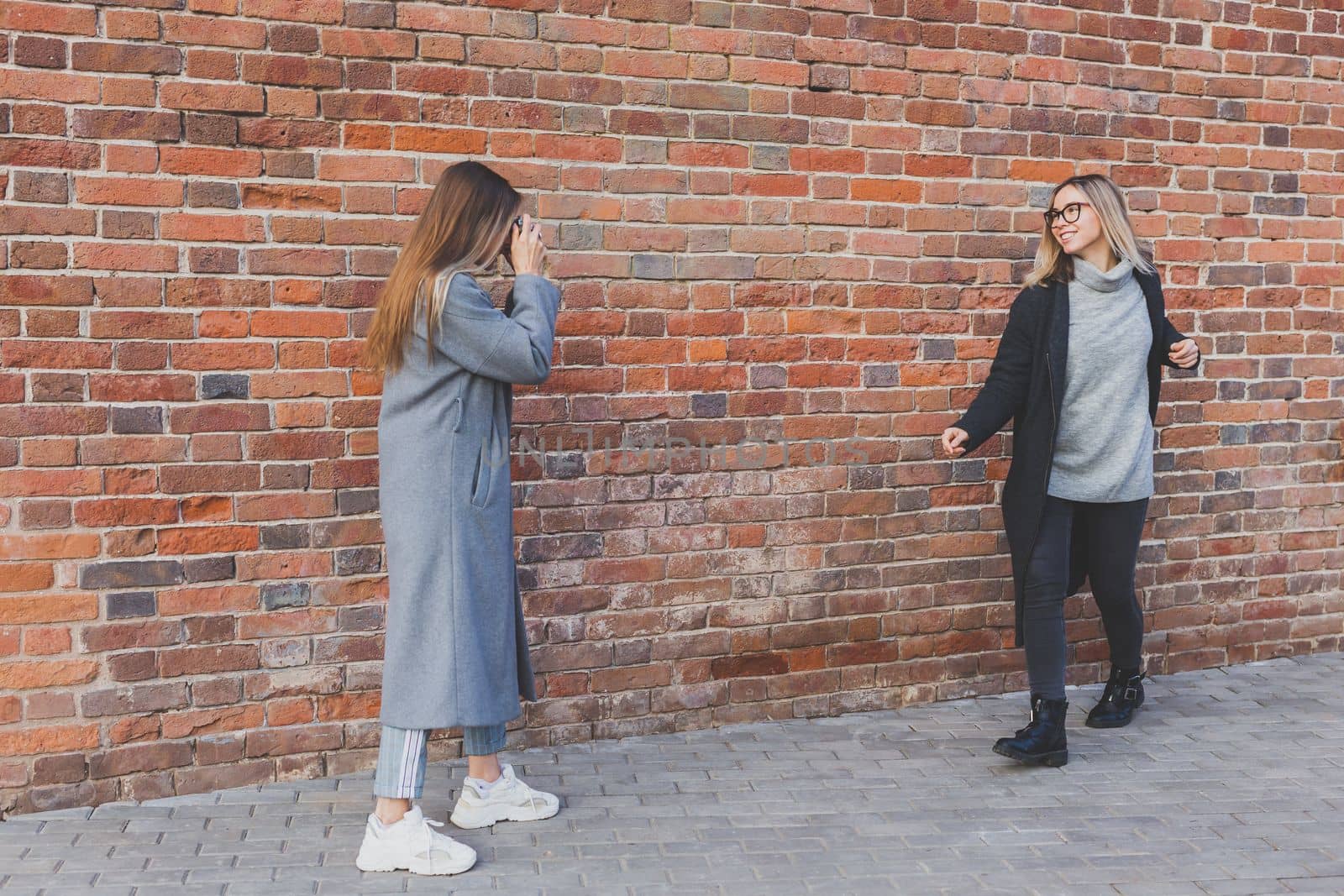 Girl takes picture of her female friend in front of brick wall in city street - photographer and vintage camera hobby concept by Satura86