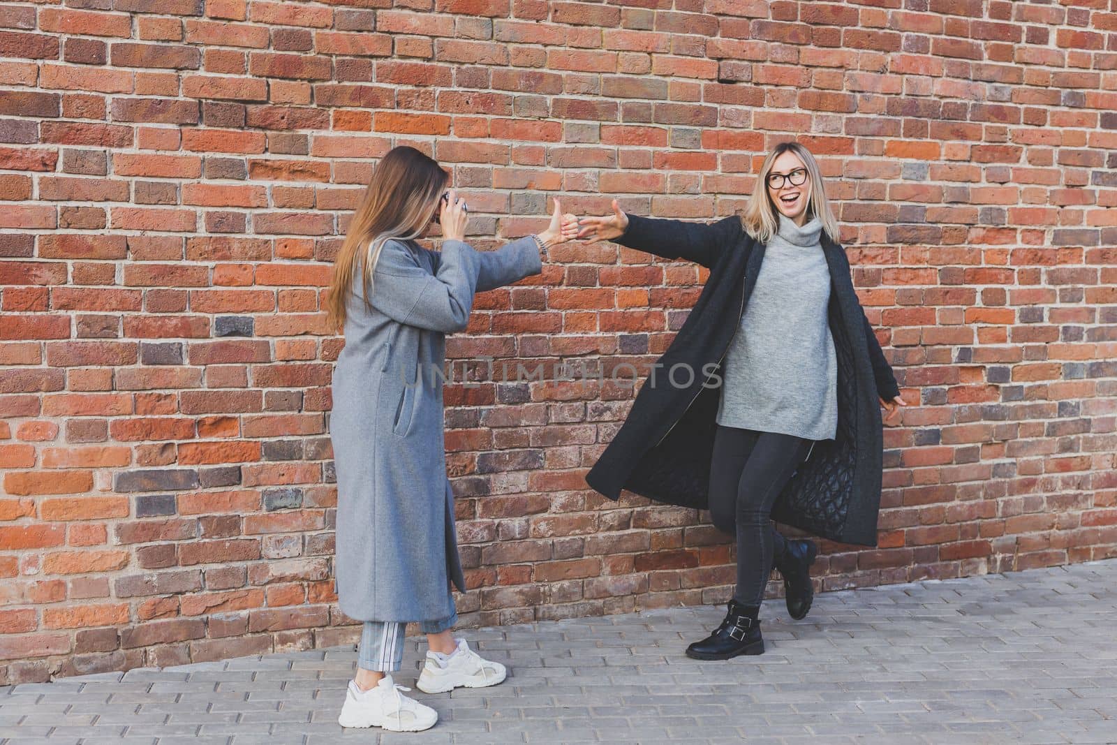 Girl takes picture of her female friend in front of brick wall in urban street - photographer and youth urban lifestyle concept by Satura86