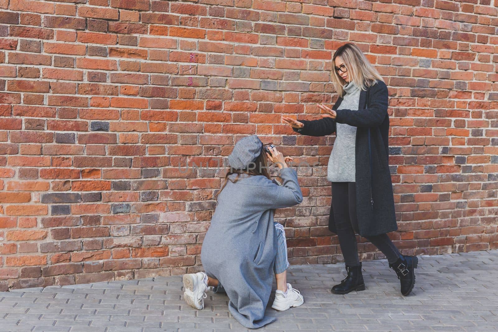 Girl takes picture of her female friend in front of brick wall in urban street - photographer and youth urban lifestyle