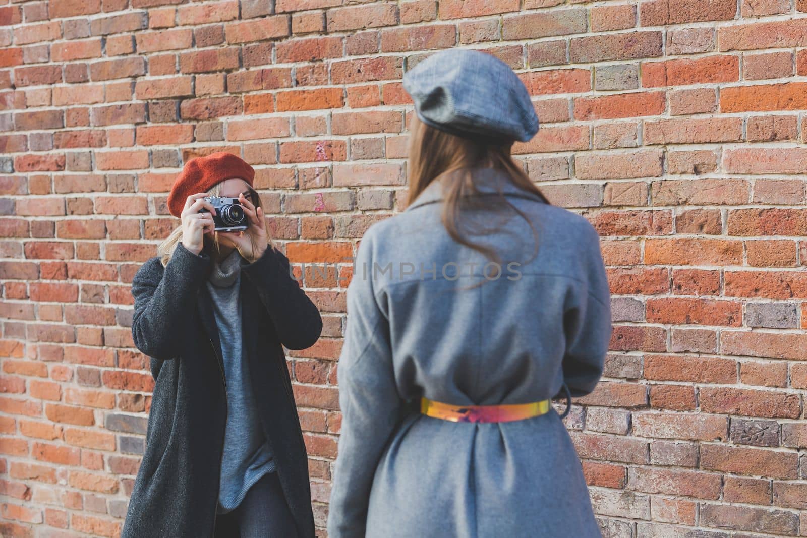 Girl takes picture of her female friend in front of brick wall in urban street - photographer and youth urban lifestyle
