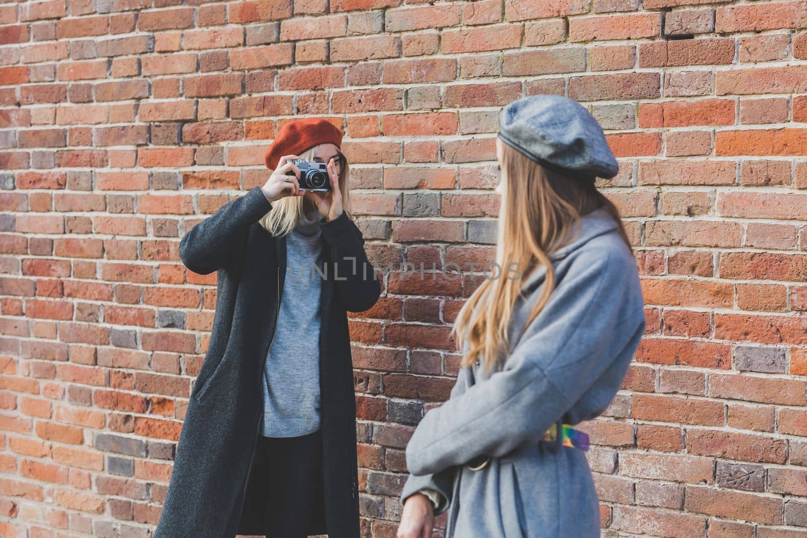Girl takes picture of her female friend in front of brick wall in urban street - photographer and youth urban lifestyle concept by Satura86